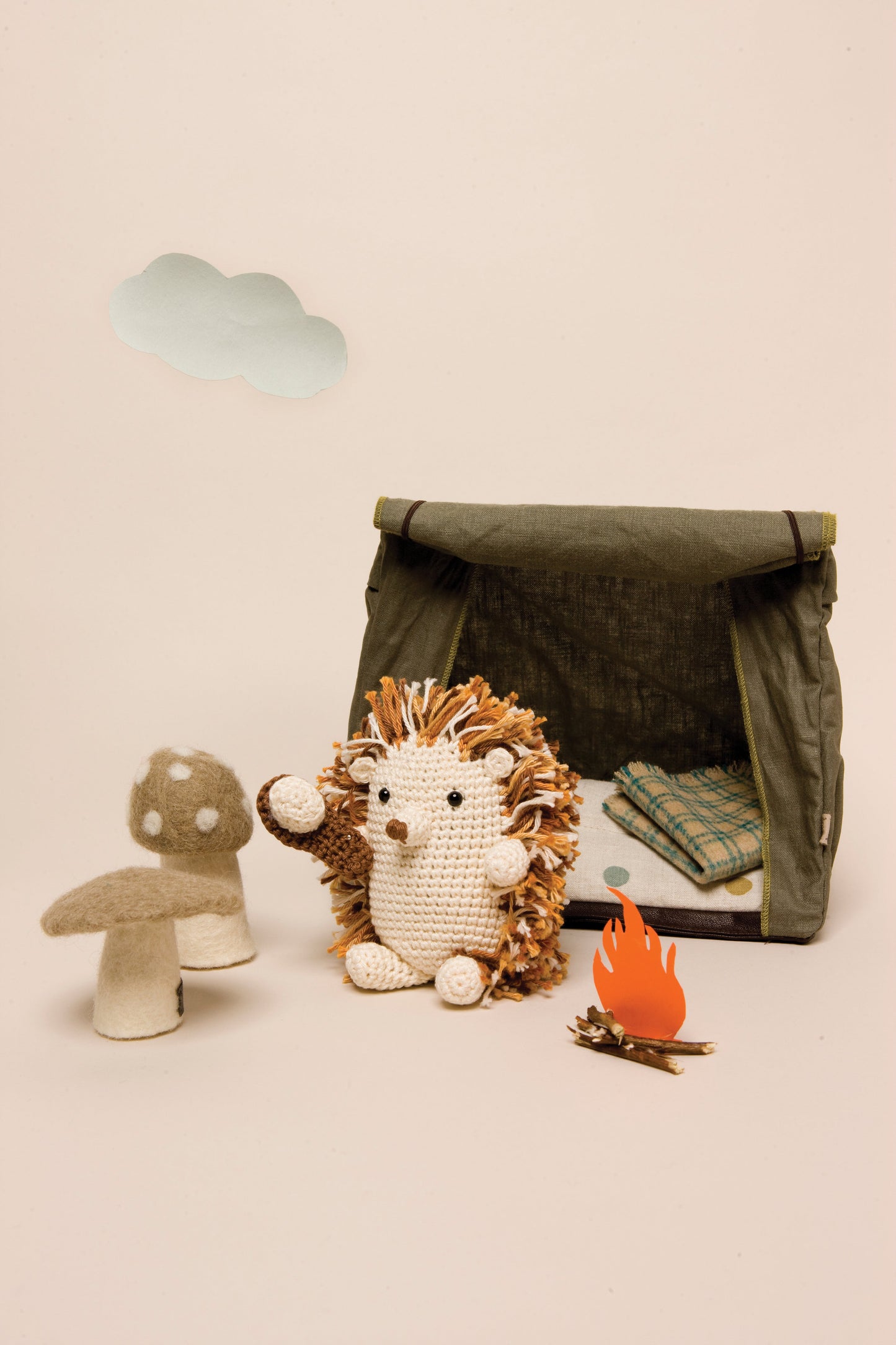 The Funny Amigurumi Digital Download from Stash Books features a crocheted hedgehog by a felt mushroom, paper campfire, and green toy tent with blanket, with a paper cloud floating against a beige backdrop.