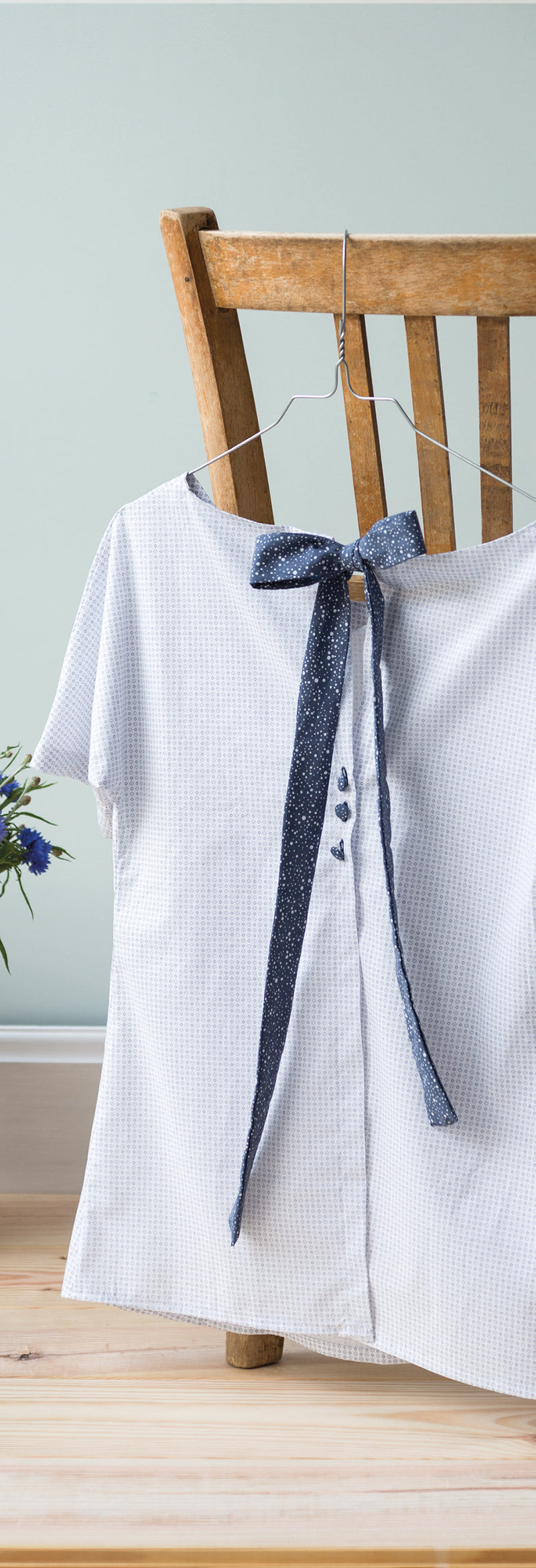 A textured, light short-sleeve blouse with a large navy bow—styled using Stash Books’ "The Complete Manual of Sewing" digital download—hangs on a chair, with a vase of blue flowers on the wooden floor nearby.
