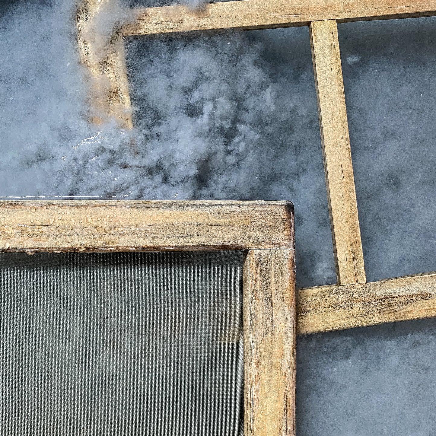 Two wooden frames for handmade paper, featured in the Stash Books Modern Papermaking Digital Download, rest diagonally over each other on a misty surface, condensation droplets visible on the mesh and wood.