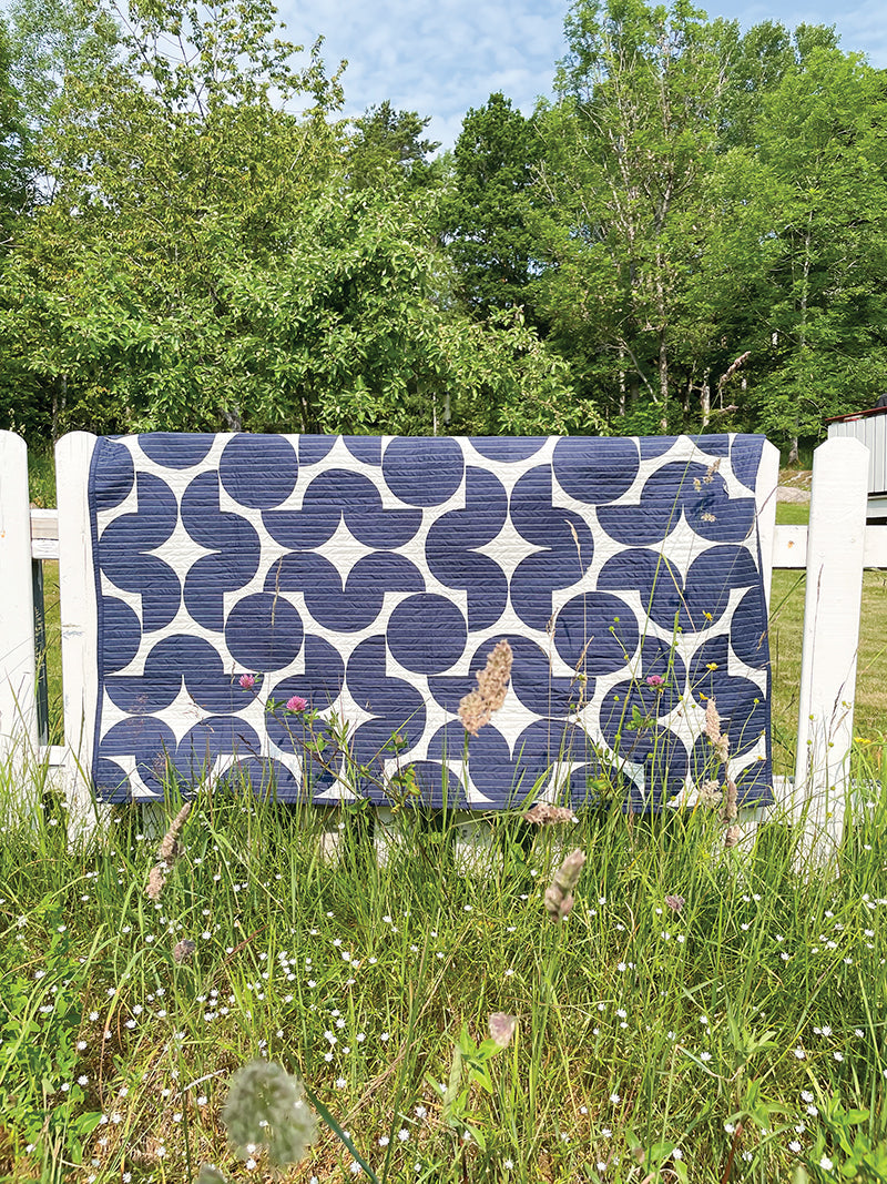 The "Quilting with Curves" digital download by Stash Books features a modern navy and white geometric quilt with circular patterns, displayed over a white fence in a grassy field beneath a partly cloudy sky.