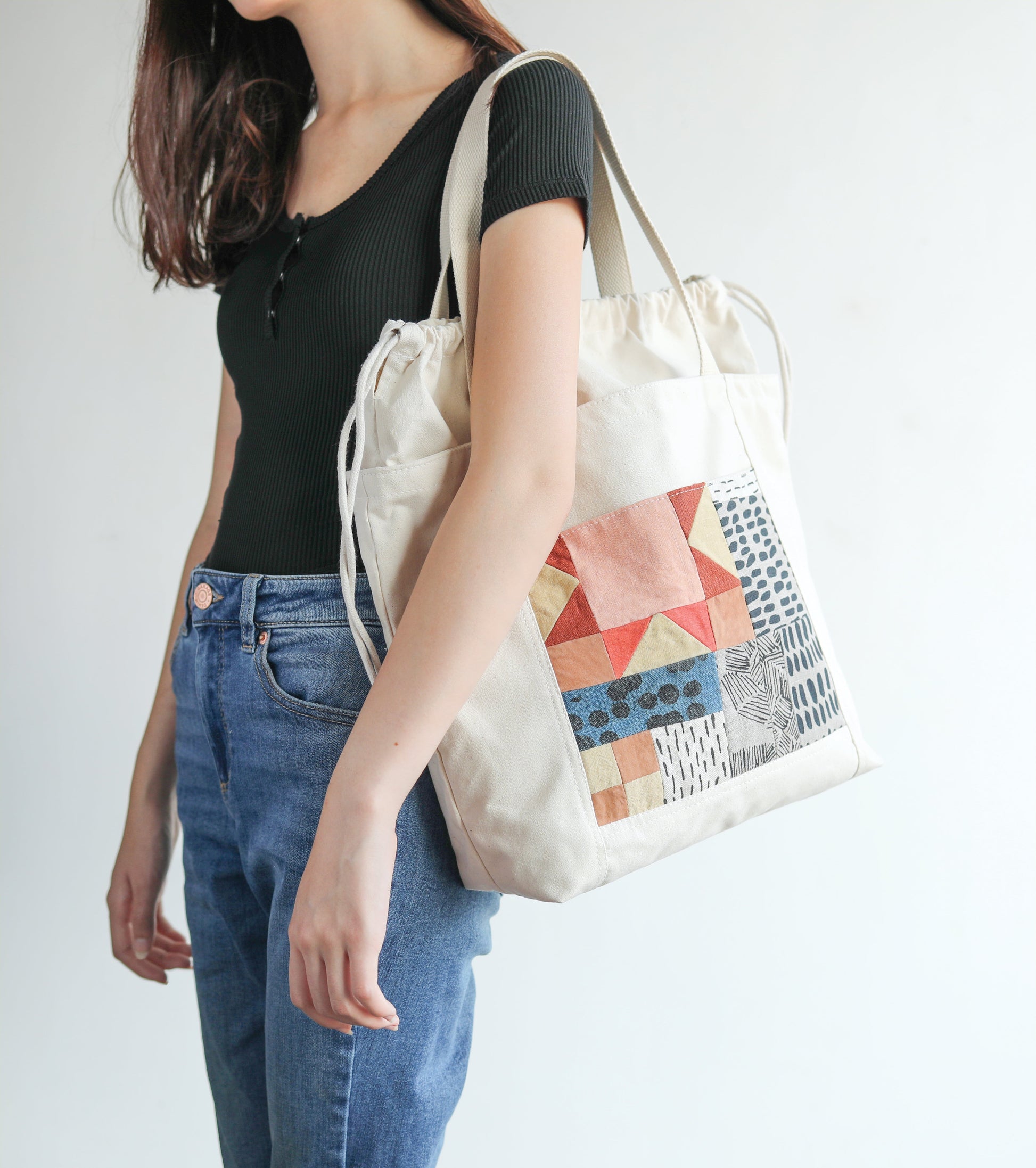 A woman in a black top and blue jeans carries the Contemporary Patchwork tote bag by Stash Books, featuring a colorful modern design, against a plain light background.