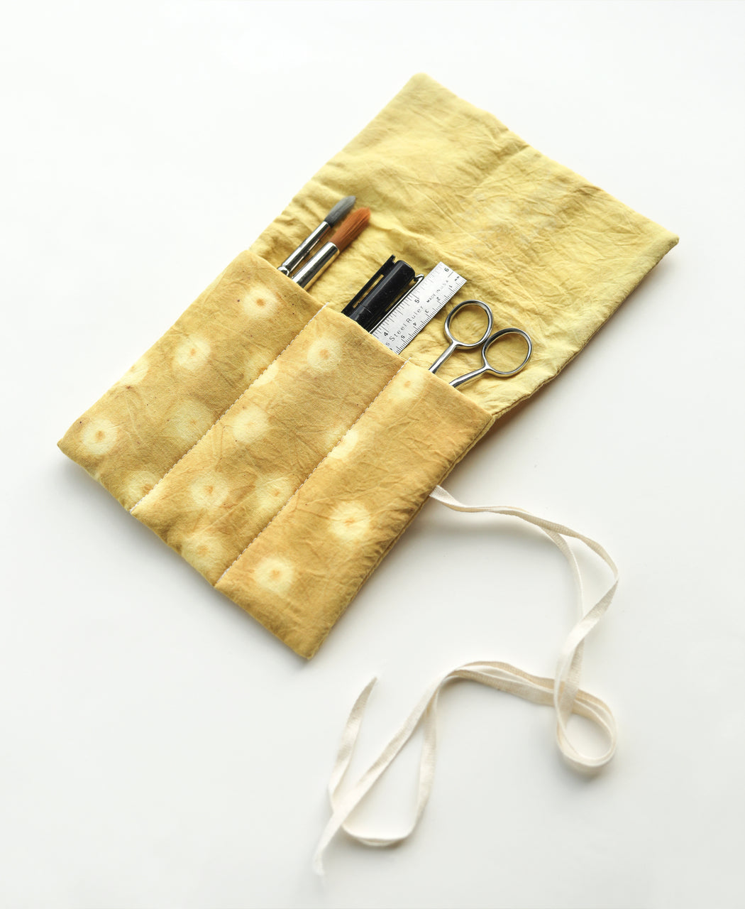 The Contemporary Patchwork pouch by Stash Books, in yellow fabric with white ties, holds art supplies such as pens, a pencil, ruler, and scissors, and is displayed on a white surface.