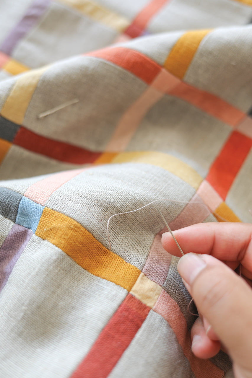 A hand stitches vibrant orange, yellow, pink, red, and blue stripes on beige fabric with a grid pattern—featuring creative surface design from "Contemporary Patchwork" by Stash Books.