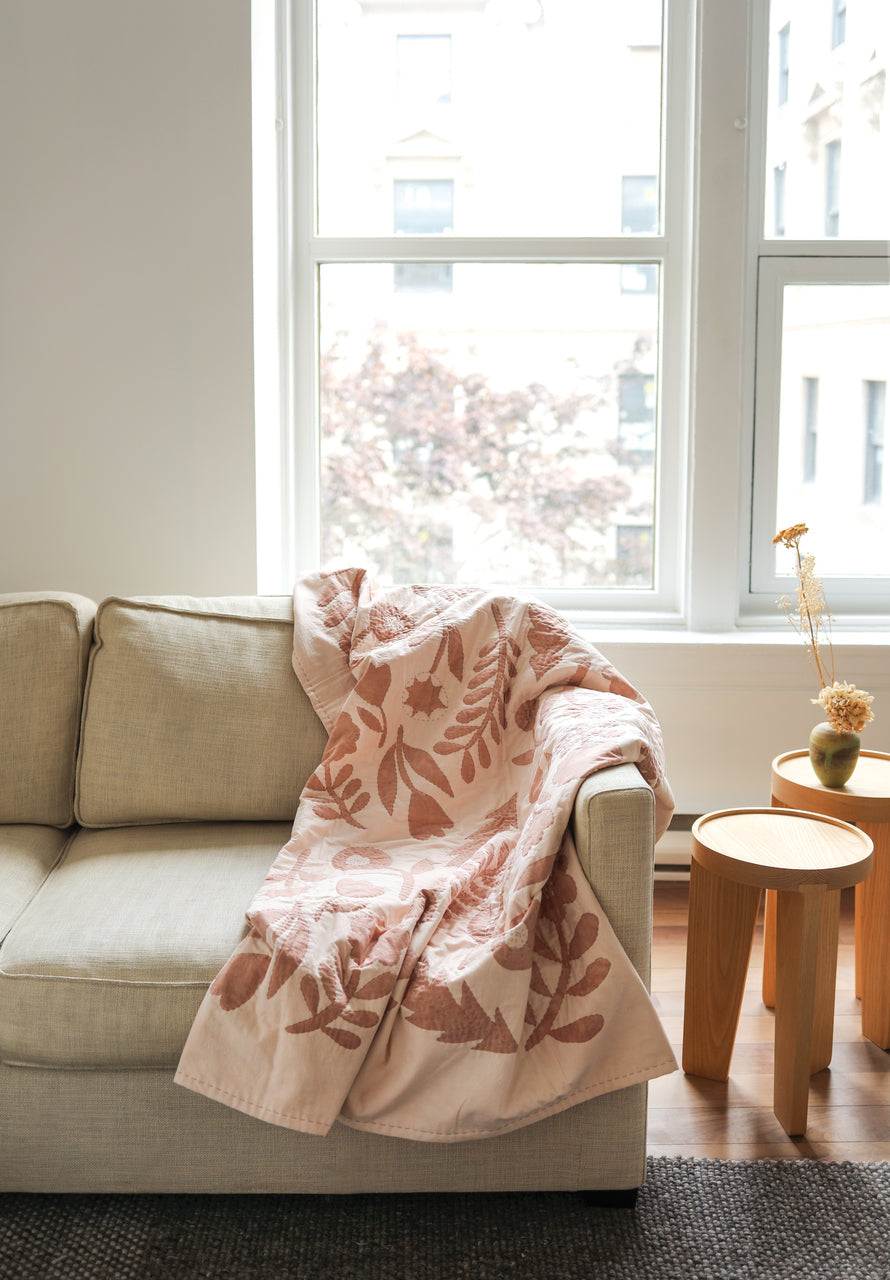 A beige sofa is accented with Stash Books' Contemporary Patchwork quilt draped over the arm, beside two wooden side tables holding a small vase of dried flowers, as daylight streams through large windows.