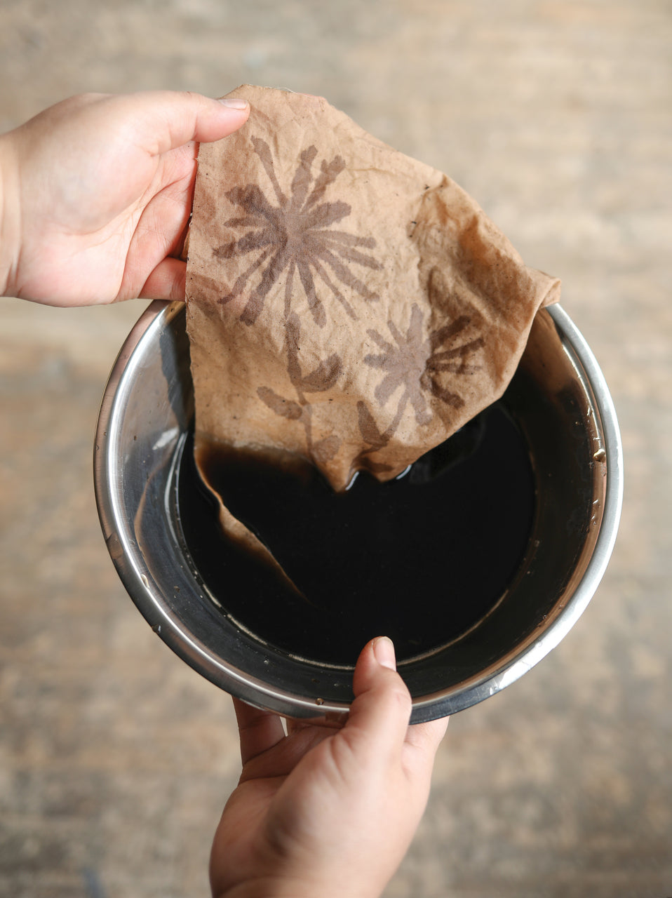 Hands hold a piece of brown paper with floral patterns above a metal bowl of dark liquid, showing a creative technique from "Contemporary Patchwork" by Stash Books, inspired by innovative textile and fabric design methods.