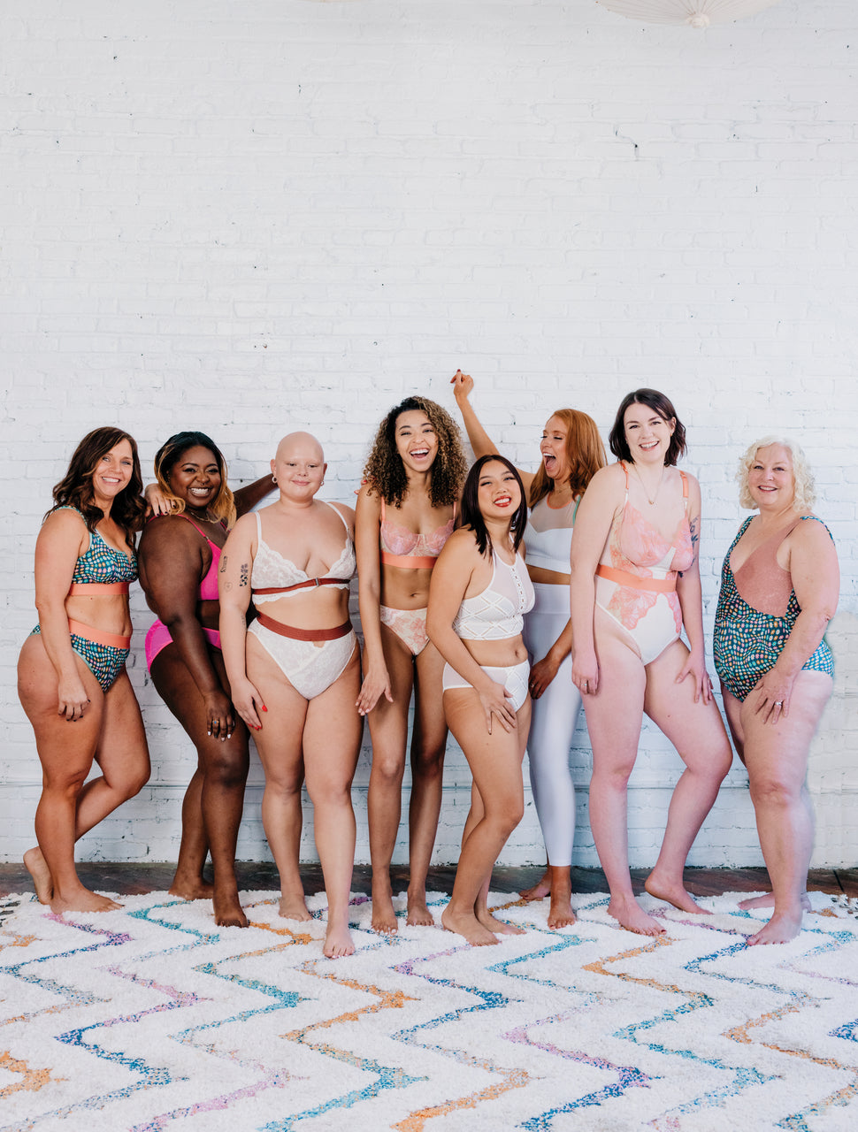 Eight women of diverse body types, ethnicities, and skin tones model lingerie made from Sew Lingerie patterns by Stash Books, smiling and laughing together against a white brick wall while standing on a vibrant patterned rug.