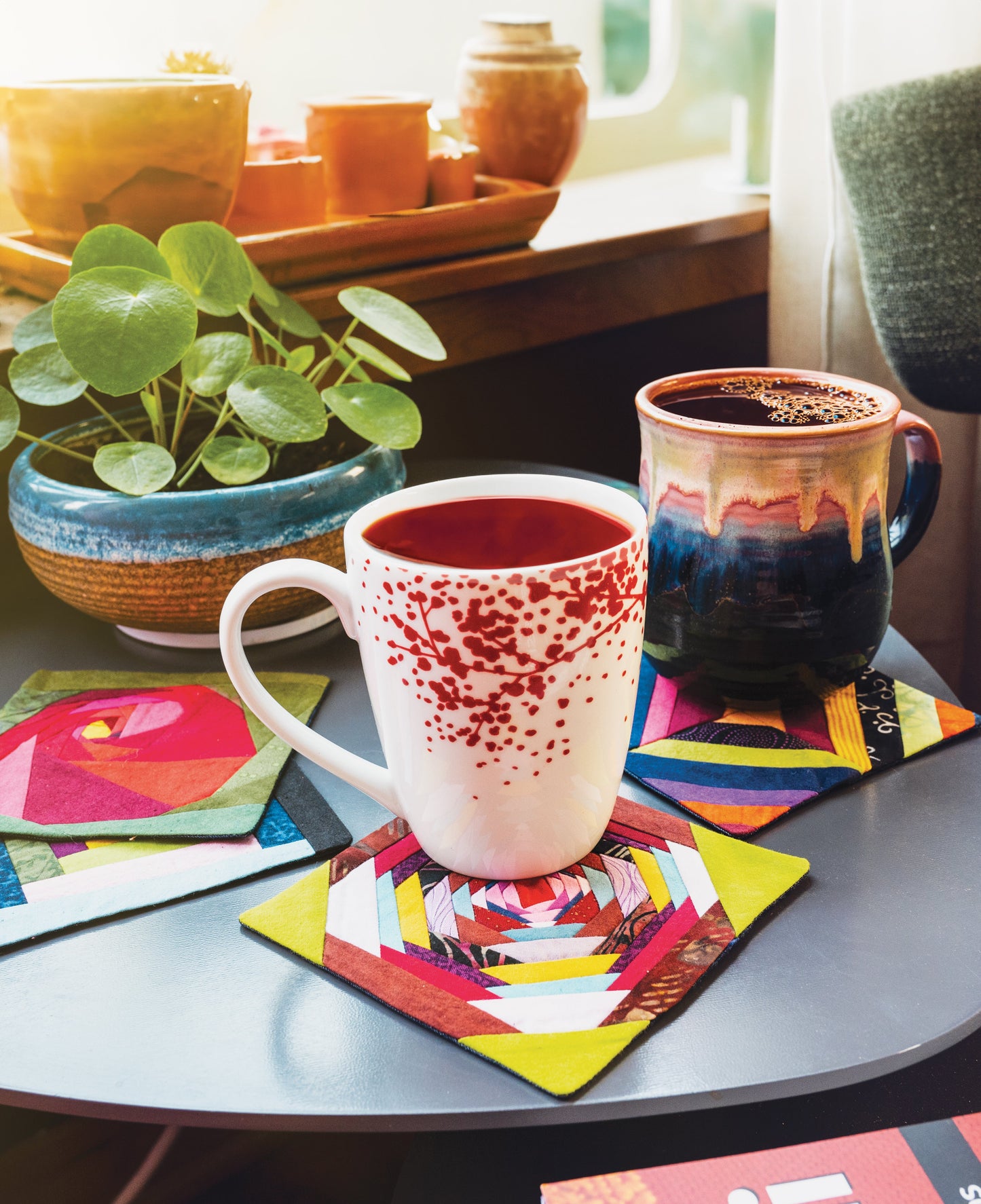 Two colorful mugs rest on a small table, each on a vibrant coaster inspired by the Scrappy Wonky Quilt Block Extravaganza Digital Download from Stash Books—a cheerful touch and great inspiration for beginner quilters.