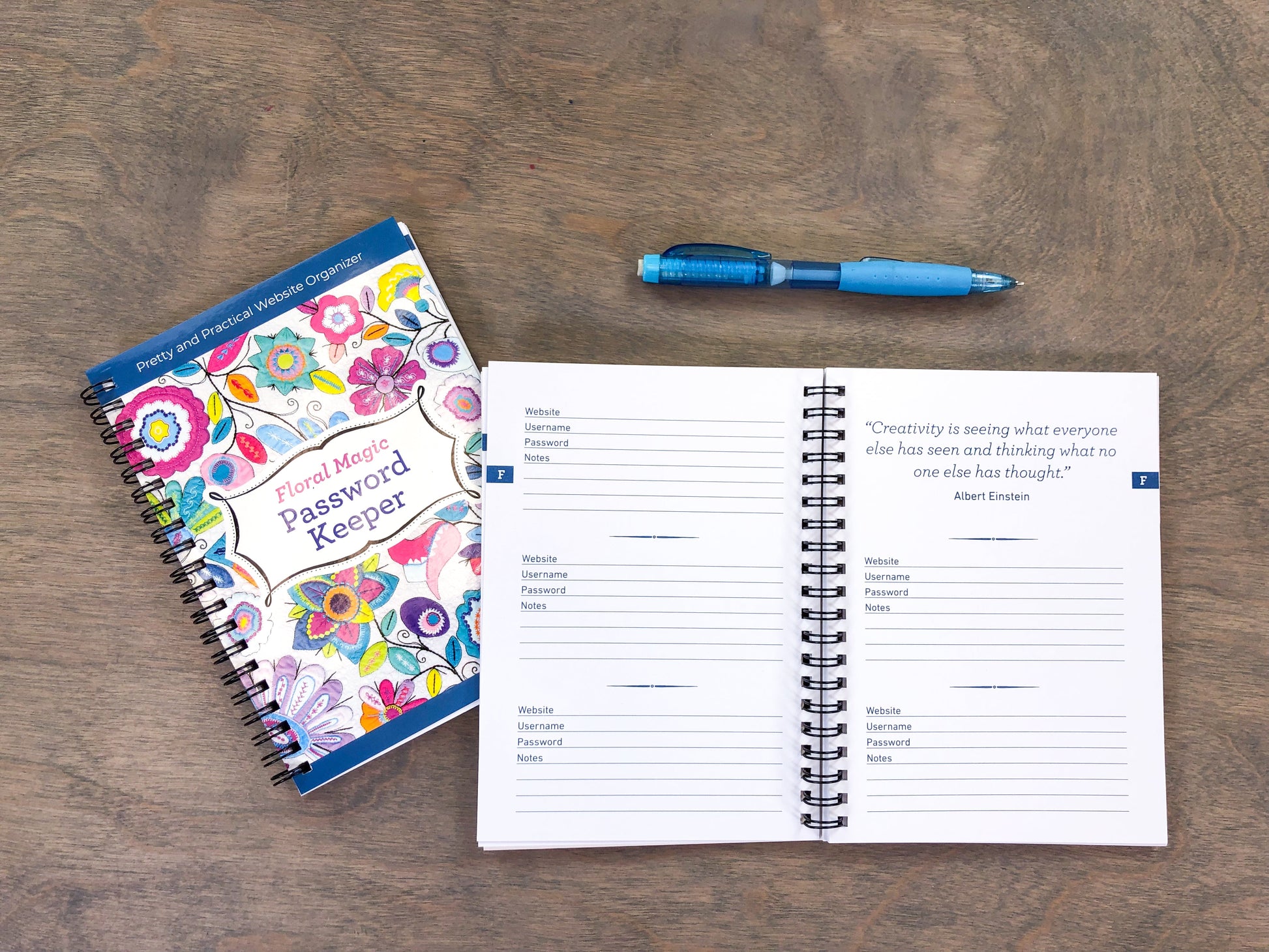 The Floral Magic Password Keeper by C&T Publishing sits open on a wooden table, displaying blank spaces for website, username, and password entries. A blue pen and the notebook's floral cover are also visible.
