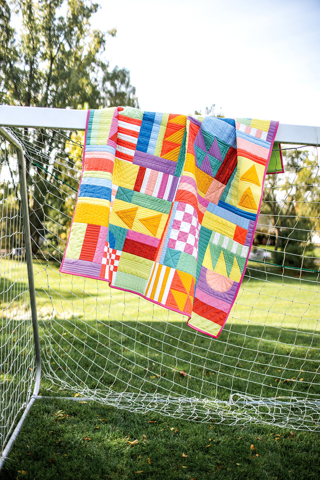 The "Modern Improv Quilting" quilt by Stash Books, showcasing beginner-friendly geometric patterns, hangs over a soccer goal on a grassy field, with trees and a clear sky in the background.