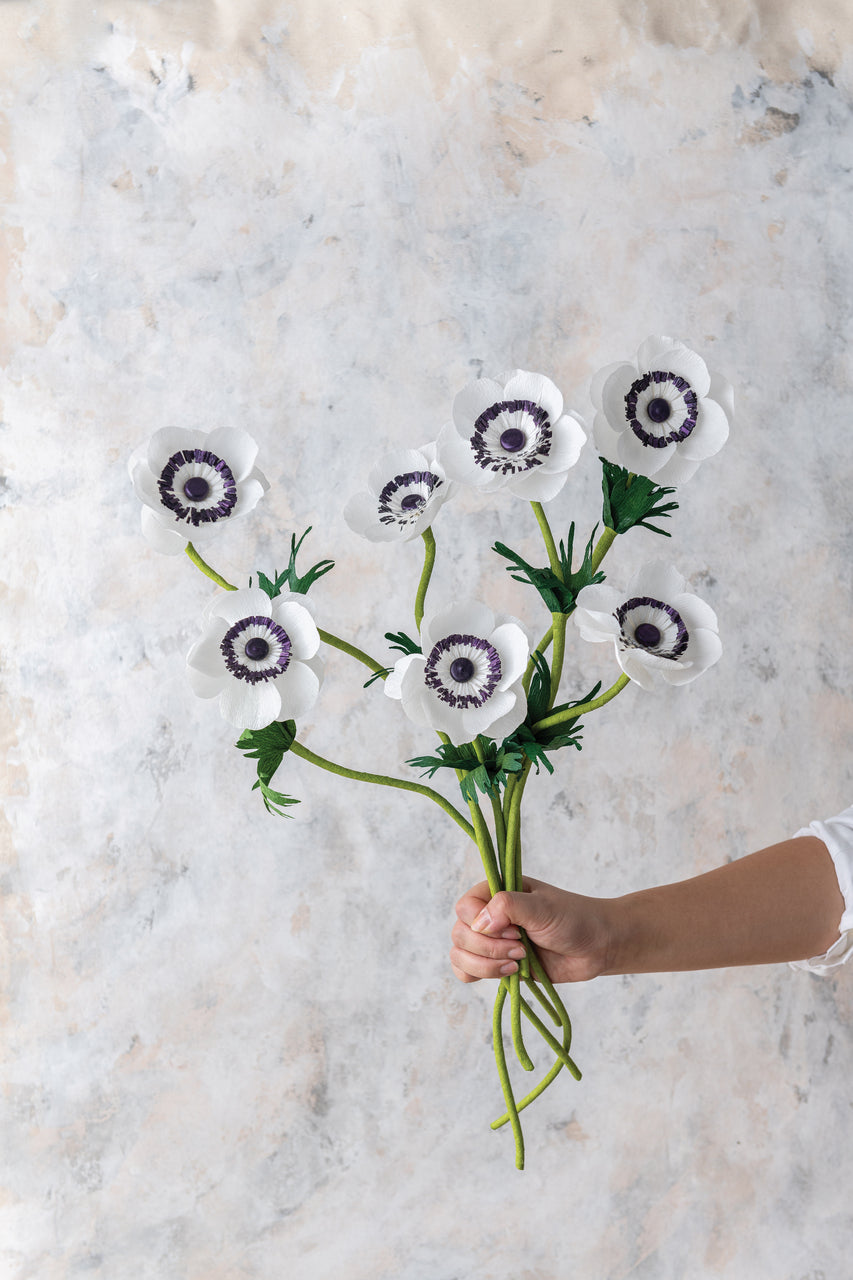 A hand holds a bouquet of white crepe paper flowers with dark centers, created using the Crafting Paper Flowers Digital Download by Stash Books, against a light, textured background.