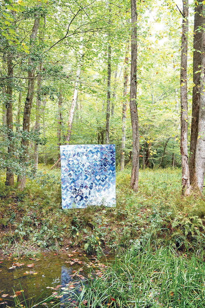 The Zero Waste Quilting Digital Download by Stash Books inspires a blue and white patchwork quilt made from leftover fabric, hanging between trees in a sunlit forest, with a tranquil pond in the foreground.