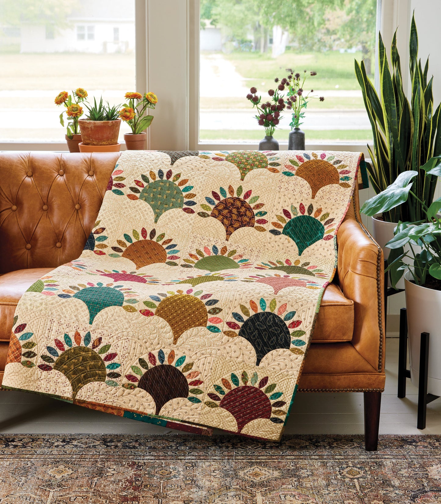 The Simple Double-Dipped Quilts Digital Download by C&T Publishing features a vibrant fan-pattern quilt draped over a brown leather sofa. Potted plants frame large windows with a yard view, and a patterned rug adds coziness to the space.