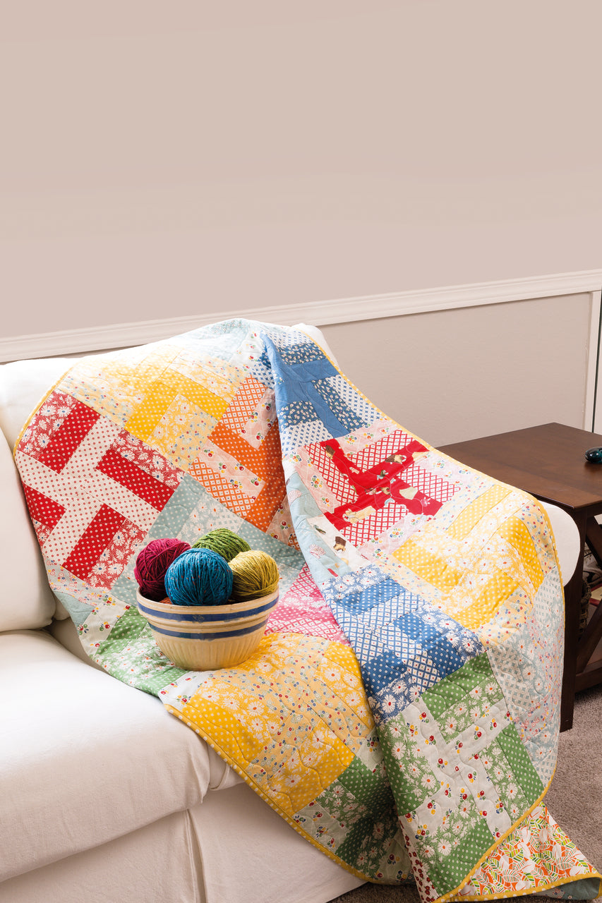 A vibrant patchwork quilt, inspired by Pat Sloan's Teach Me to Make My First Quilt Digital Download from C&T Publishing, is draped over a white sofa. On top sits a bowl of green, red, blue, and yellow yarn balls. A wooden side table is nearby.