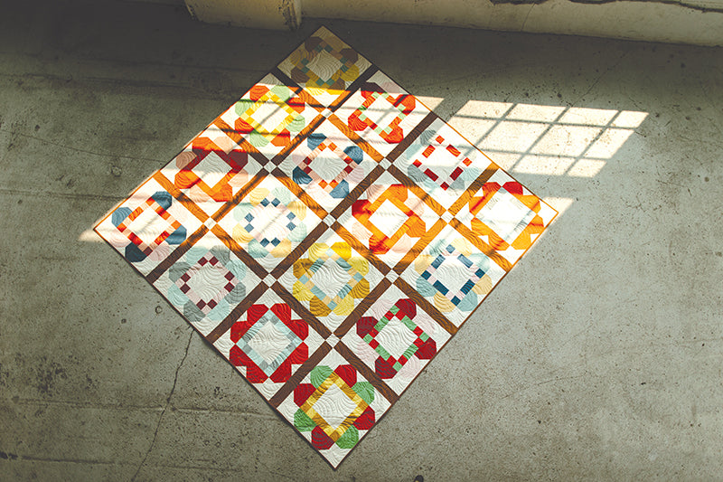 The Patchwork Companion by Stash Books lies on a concrete floor, sunlight streaming through a window and casting grid-like shadows across its colorful geometric quilt patterns.
