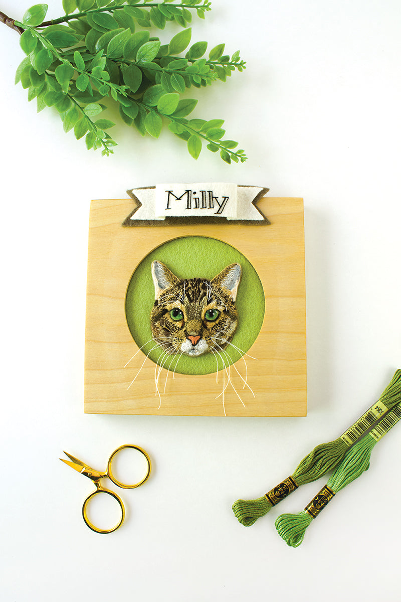A wooden frame displays an embroidered tabby cat portrait labeled Milly, made with thread painting on a green background. Nearby are gold scissors, green embroidery threads, and plant leaves. Product: Felt & Thread by Stash Books.