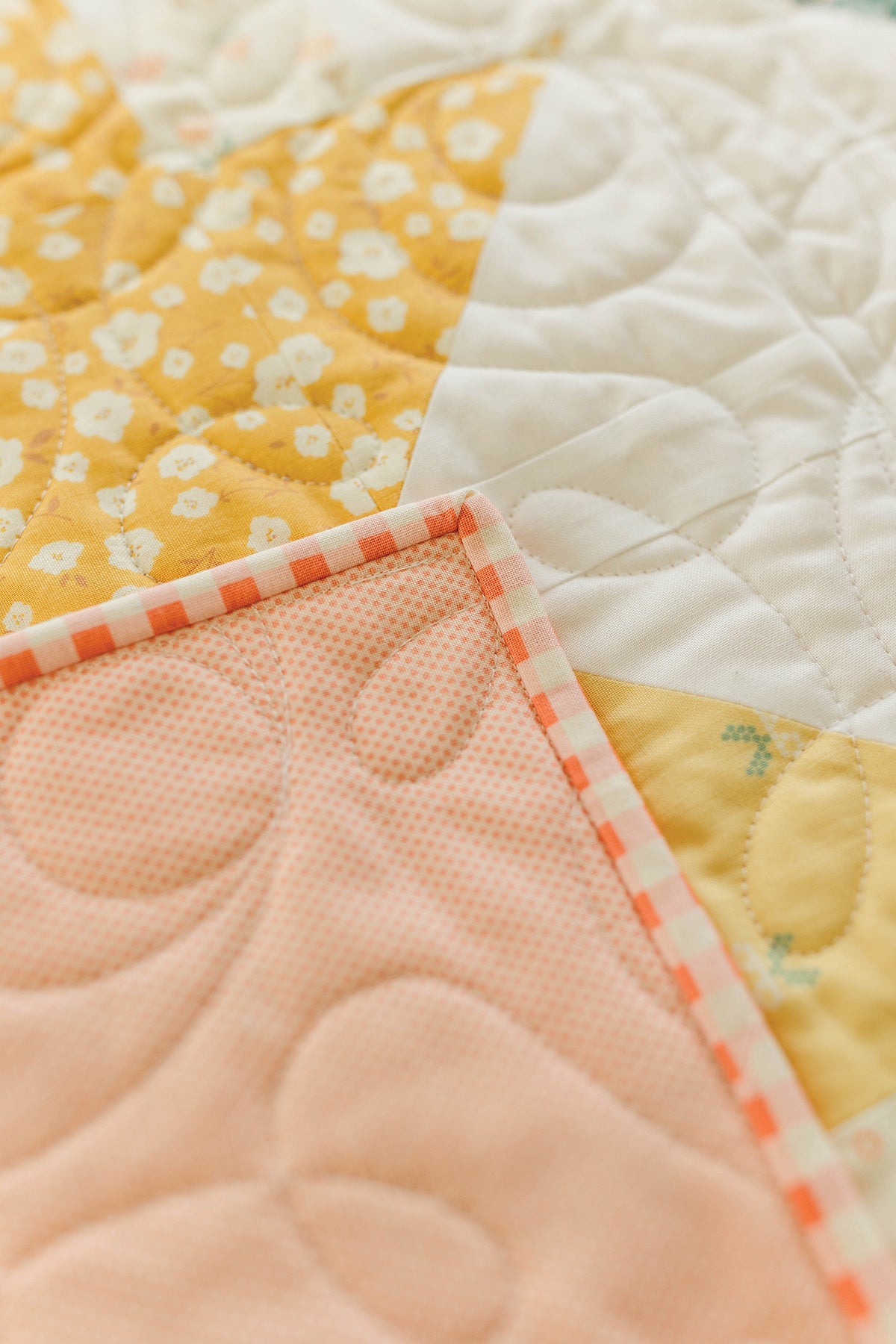 A close-up of Layered & Stacked by Stash Books—a quilt in yellow floral, white, and peach fabrics with pink and white checkered binding and swirled quilting—ideal inspiration for beginner quilting or new quilt designs.