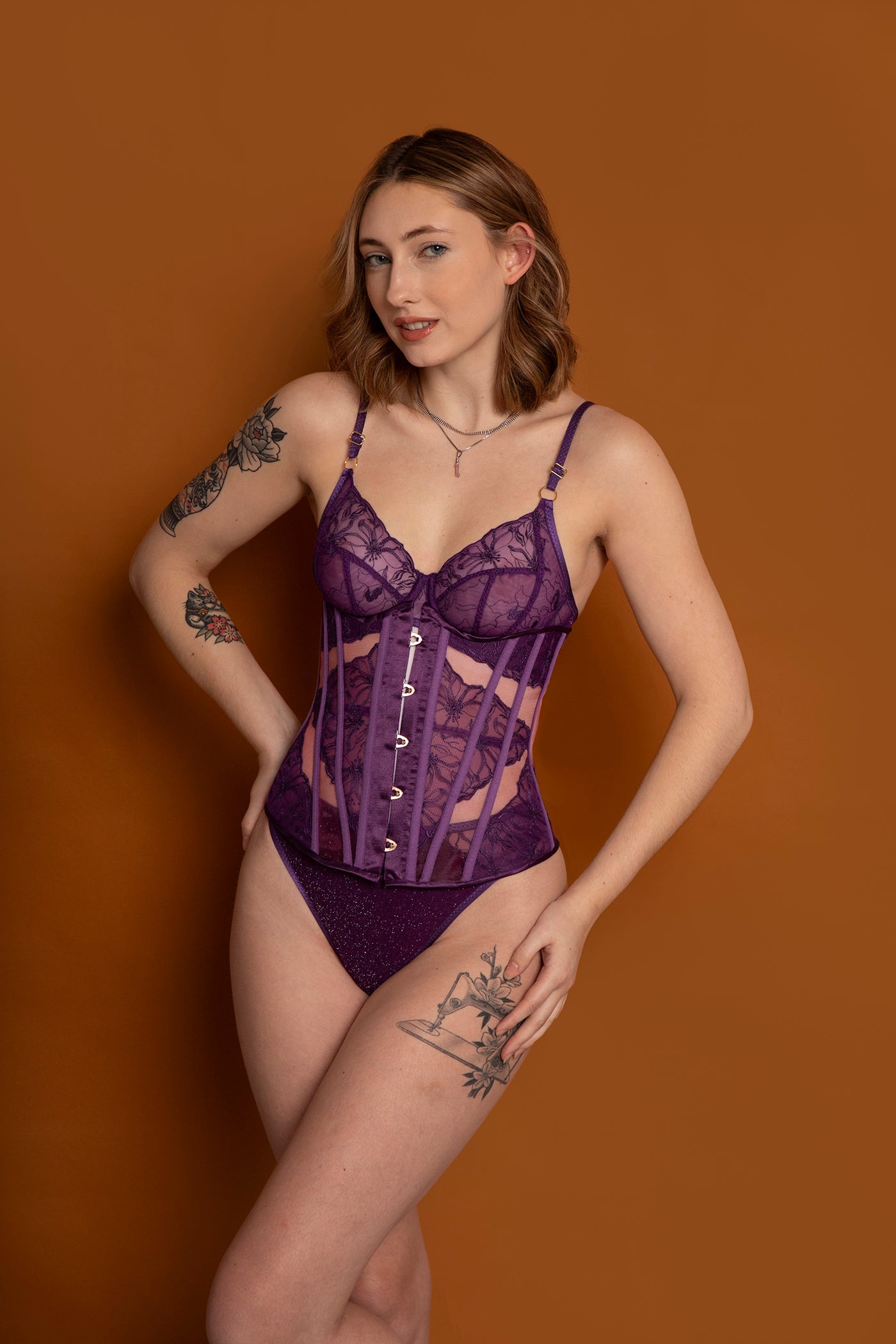 A woman with shoulder-length hair and tattoos poses confidently against a brown background, modeling expertly constructed purple lingerie inspired by "Sew Underwire Bras & Corsets" from Stash Books.