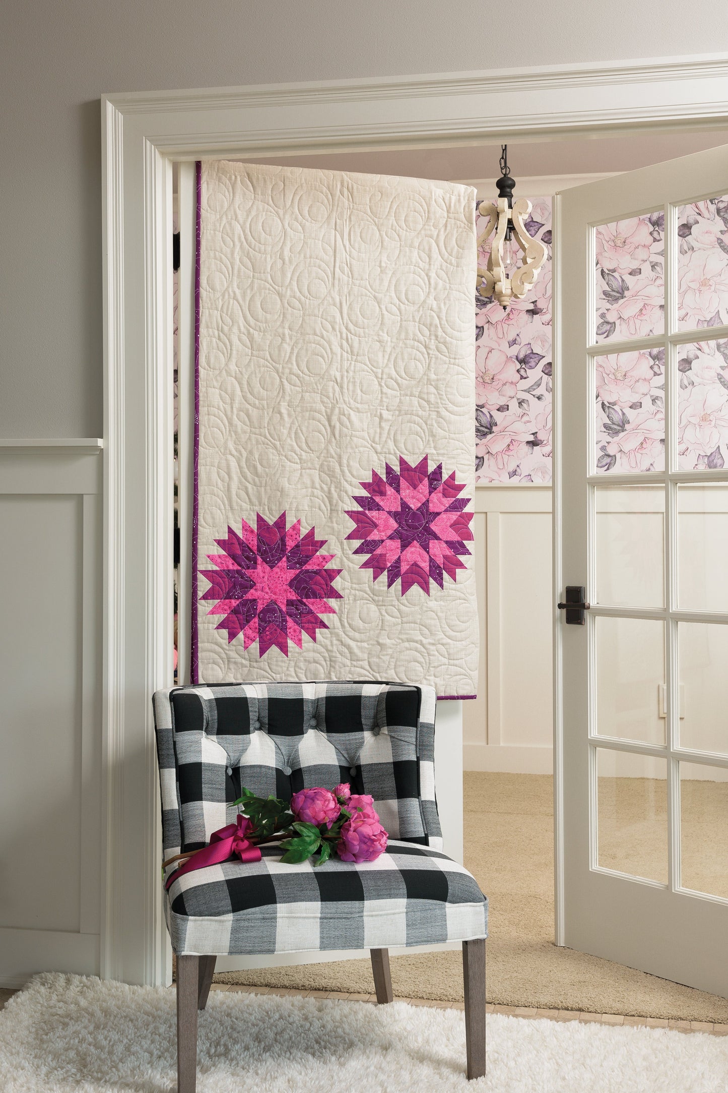 A black and white plaid chair with pink flowers sits by a doorway. Hanging in the doorway is a quilt made using the Spectacular Stars Simplified Digital Download from C&T Publishing, with floral wallpaper seen in the next room.