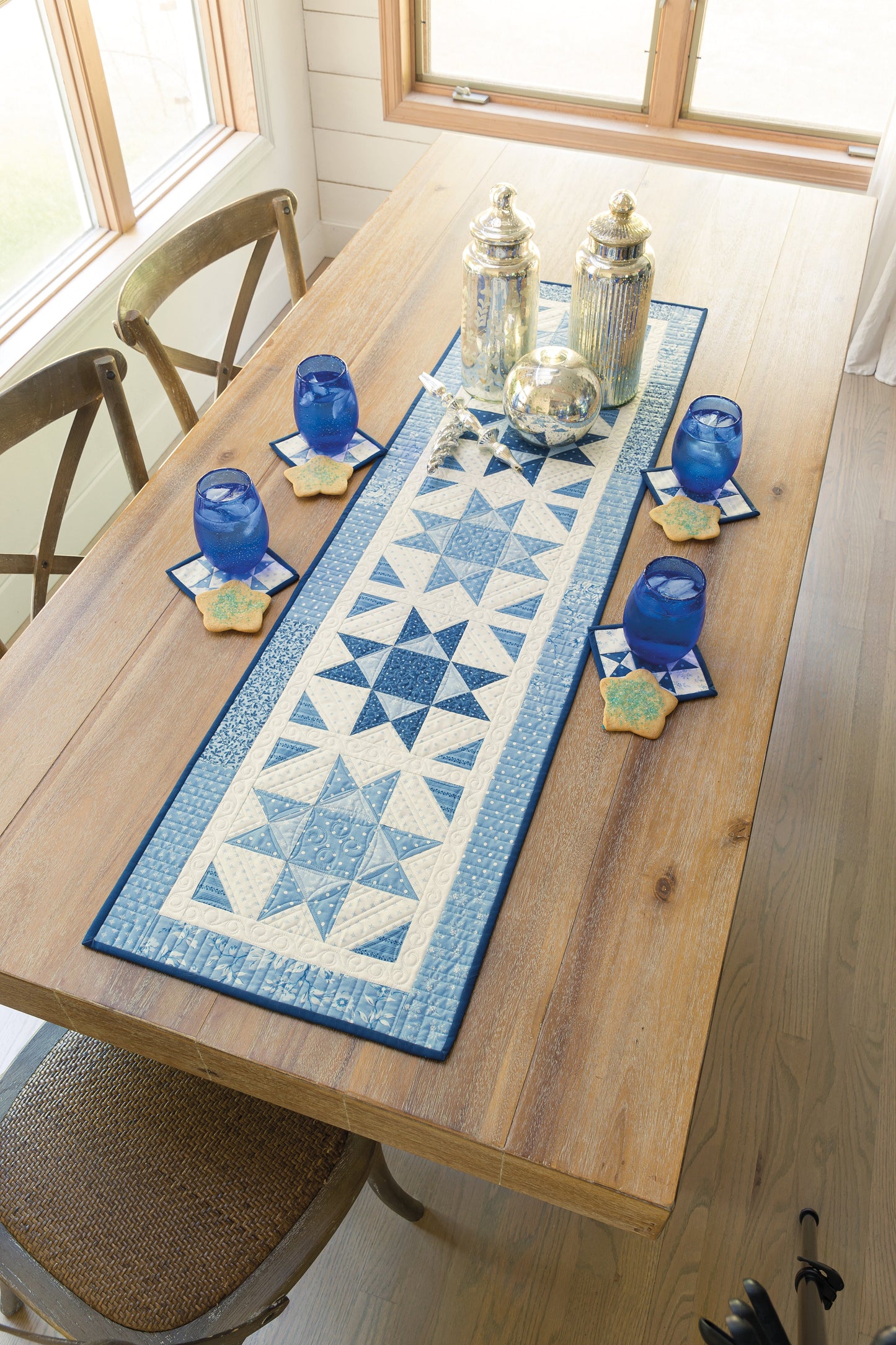 In a sunlit room, a wooden dining table features the "Home for the Holidays Digital Download" by C&T Publishing—a blue and white geometric quilted table runner, four blue glasses on star coasters, and three silver jars with wooden chairs.