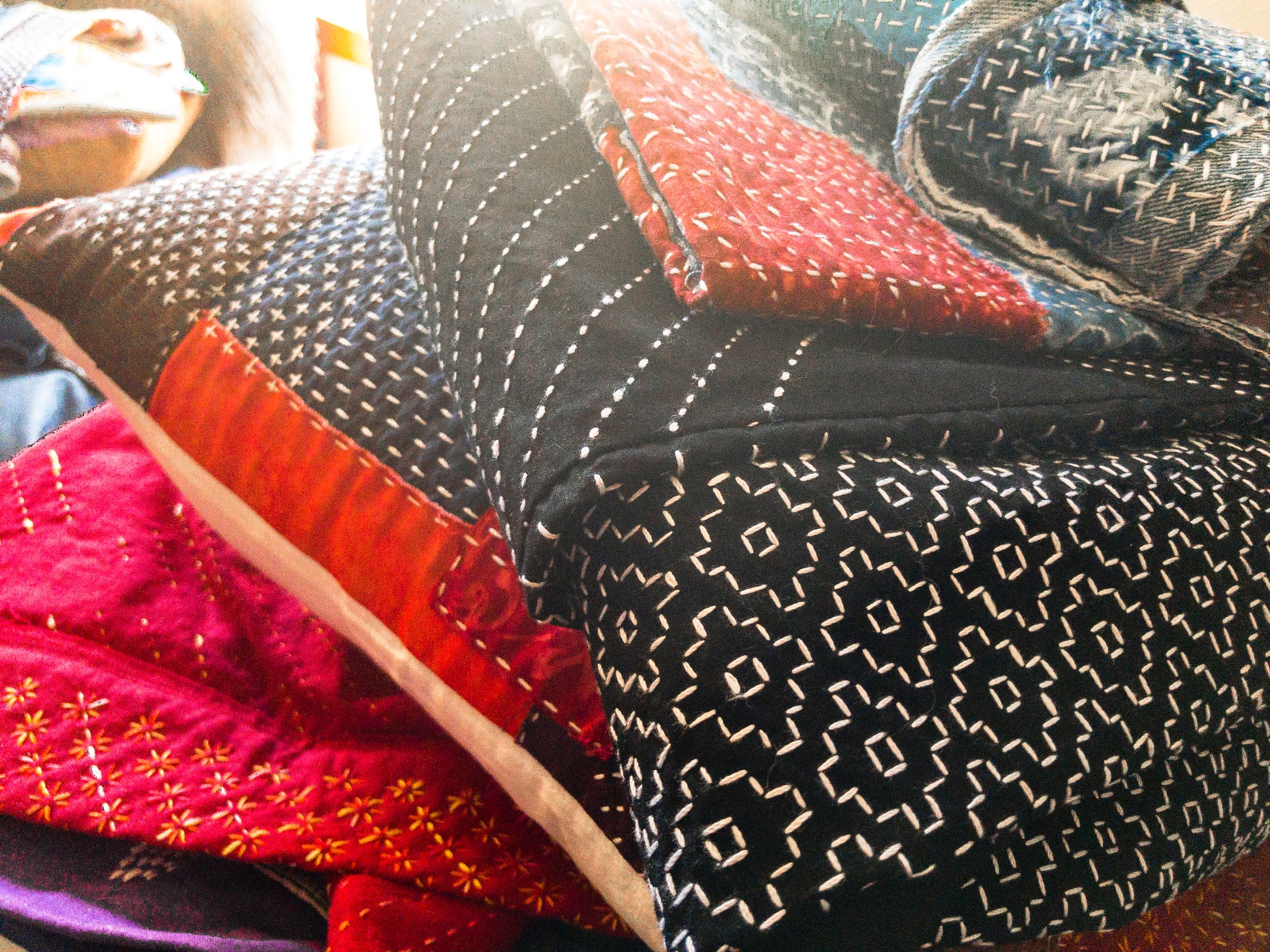 A close-up of Sashiko 101: Iconic First Stitches by Creative Spark shows hand-stitched quilts with geometric patterns in black, white, red, and blue fabrics, highlighting delicate Sashiko stitches and expert craftsmanship in natural light.