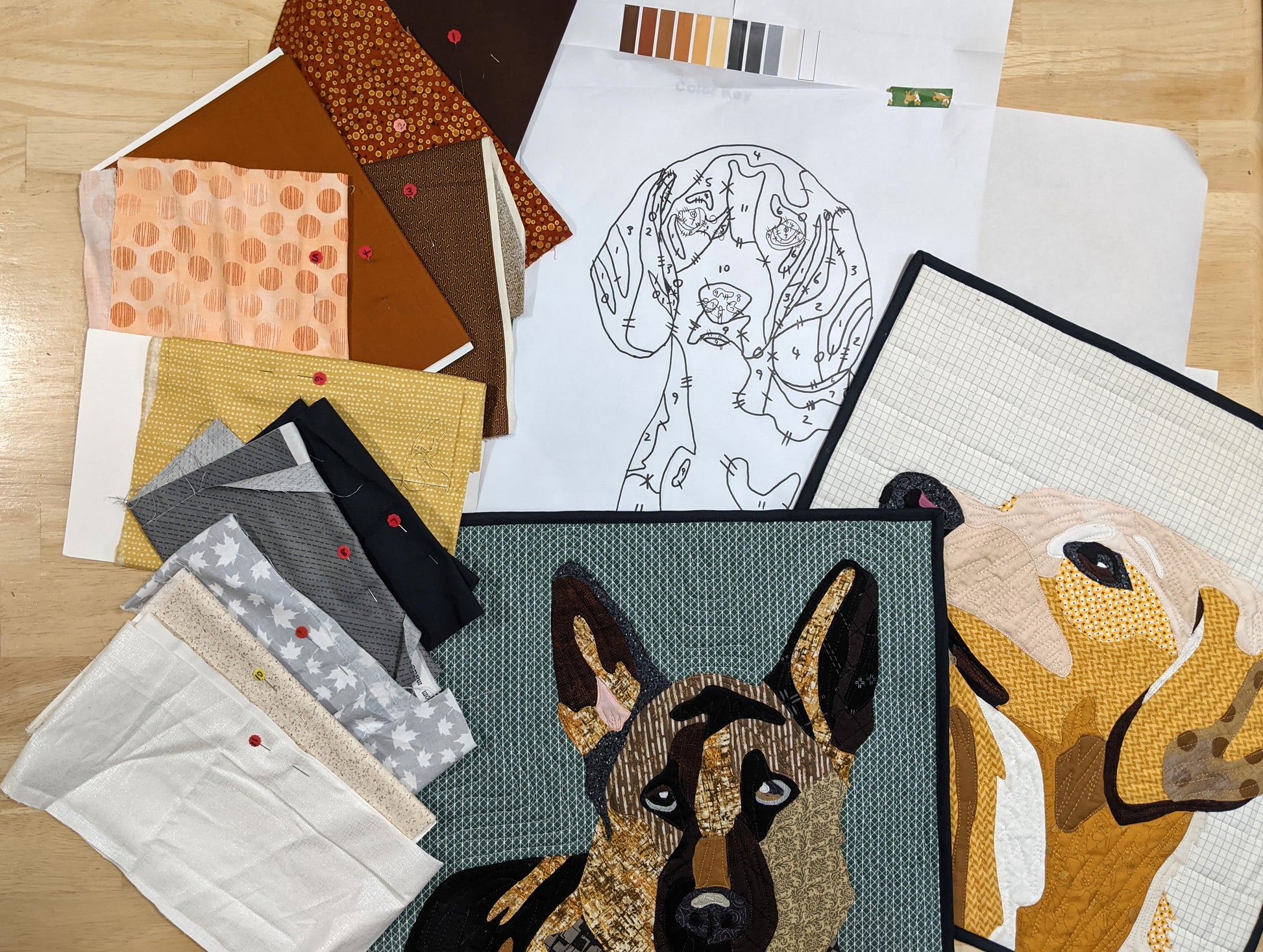 A workspace with fabric pieces, quilted dog portrait mini quilts, a dog line drawing, color palette, and sewing pins on wood showcases a Paint By Number Quilts: Dog Portraits project by Creative Spark using turned edge appliqué.