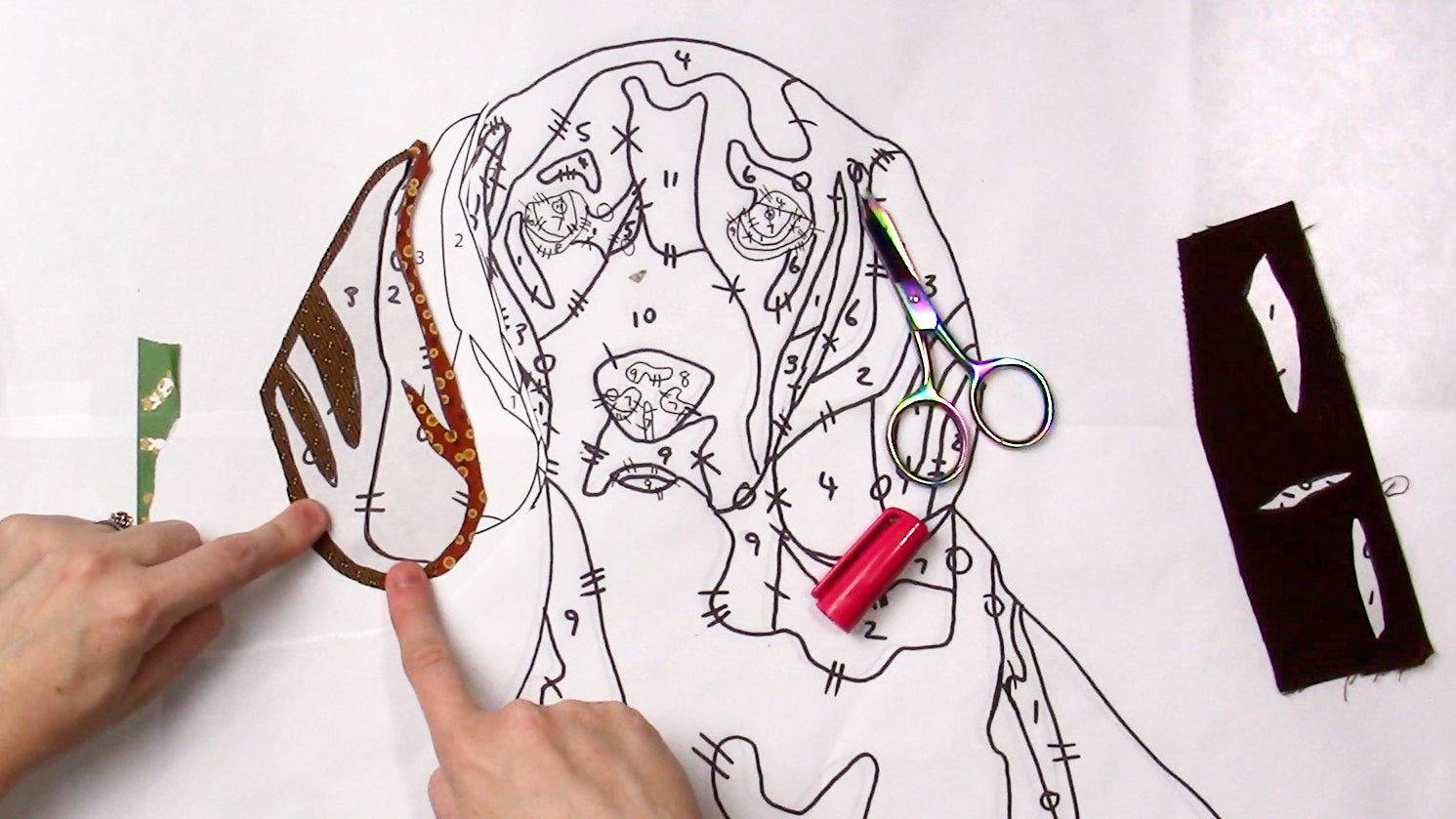 A person points to a numbered dog outline on white paper with fabric pieces, scissors, and a pink marker—showcasing Creative Spark's "Paint By Number Quilts: Dog Portraits" kit for turned edge appliqué.