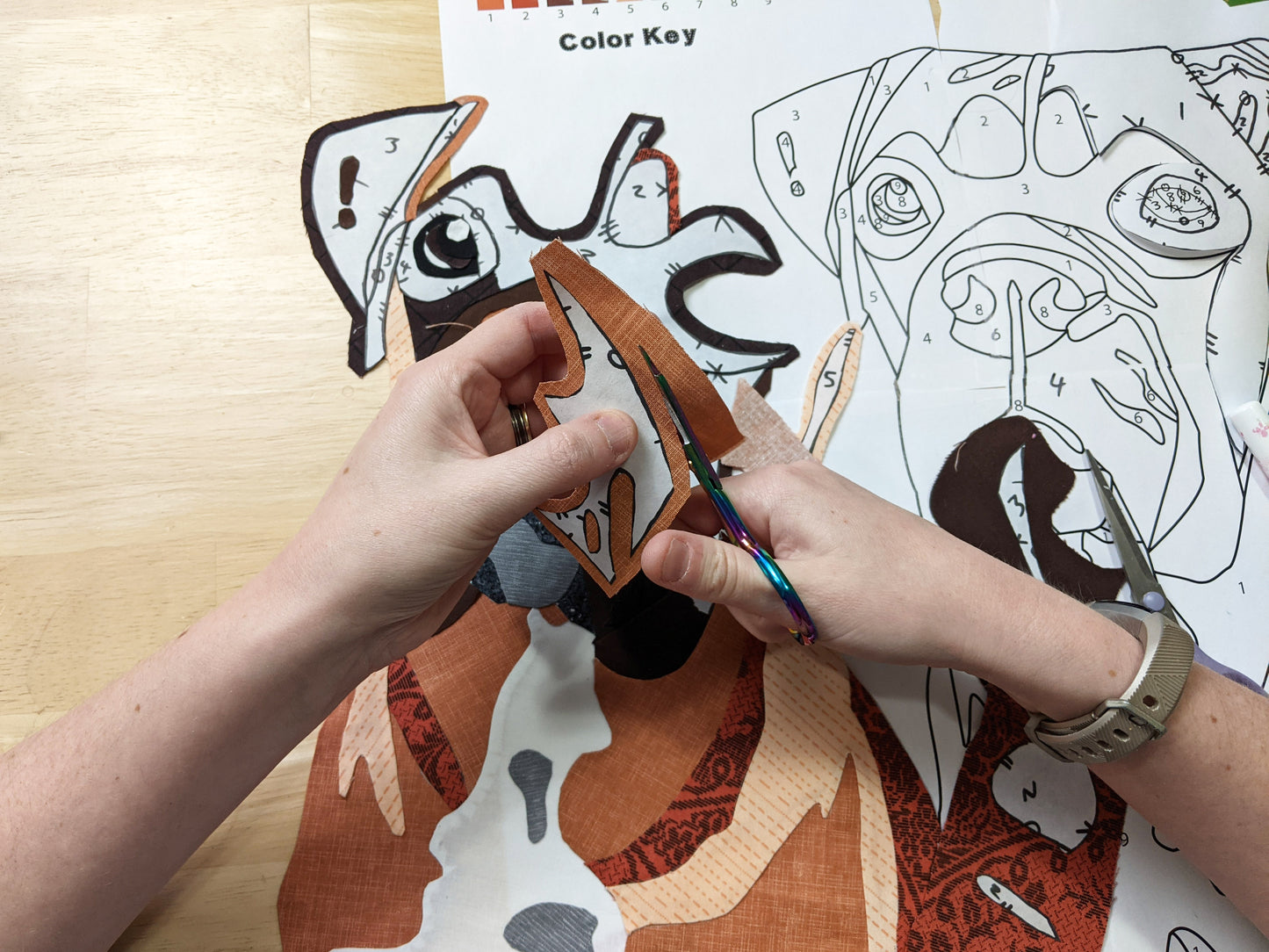 A person uses Creative Spark's Paint By Number Quilts: Dog Portraits to cut brown fabric by a color key and pattern, using freezer paper for accuracy, while crafting an animal quilt project on a wooden table.