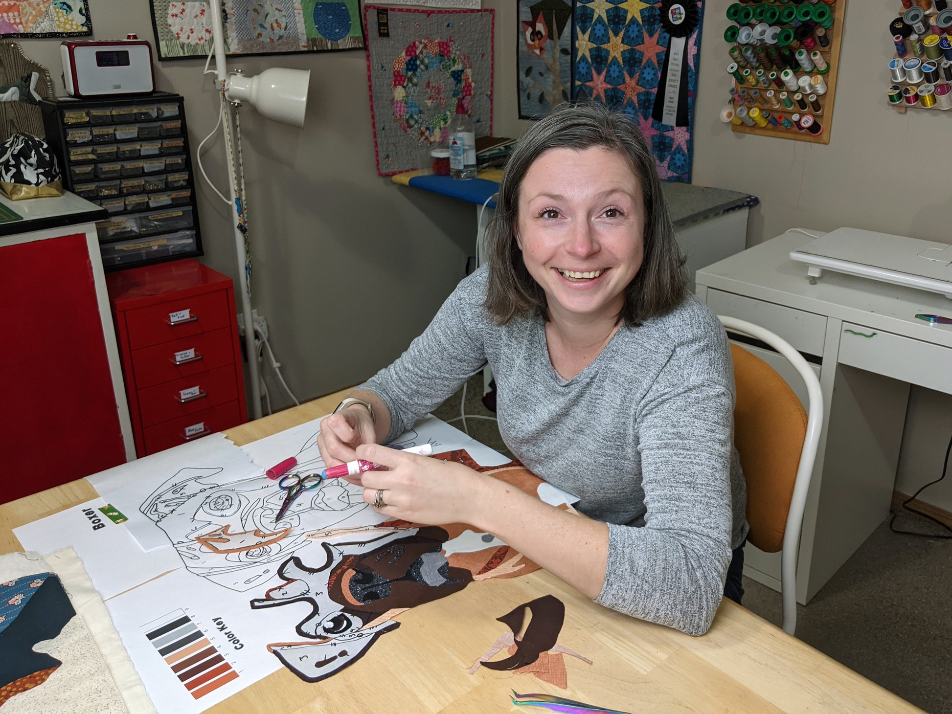 A woman with shoulder-length brown hair smiles as she works on the "Paint By Number Quilts: Dog Portraits" kit by Creative Spark at a craft table, surrounded by sewing supplies and colorful decor.