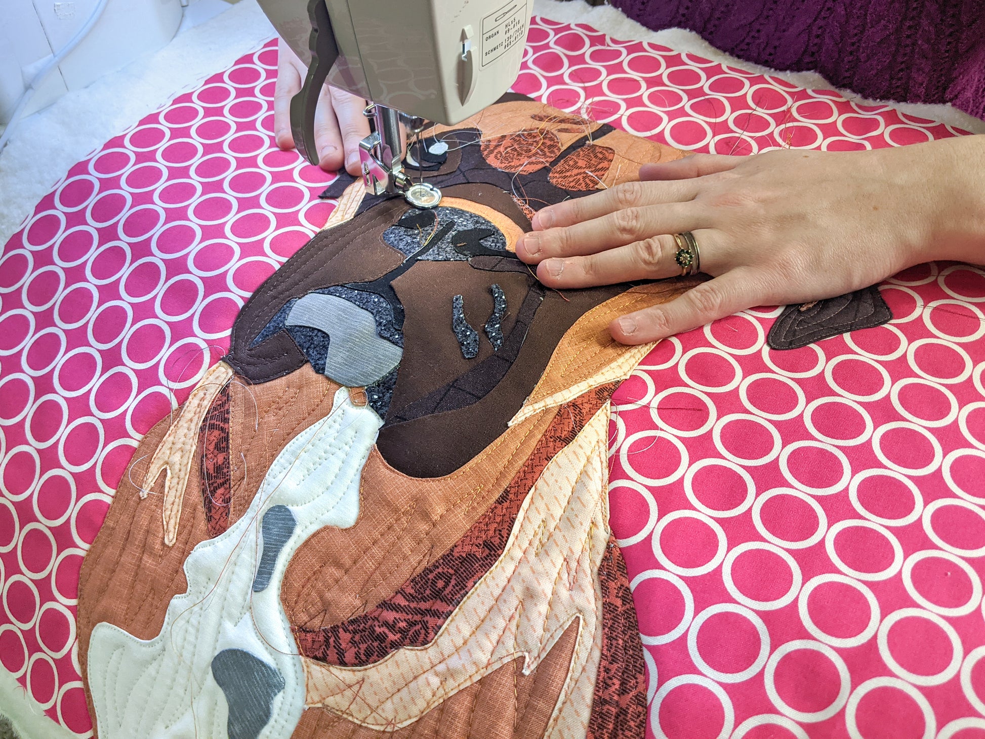 A person uses the Creative Spark Paint By Number Quilts: Dog Portraits kit to quilt a vibrant dog design on pink and white polka dot fabric, guiding the material under the sewing machine needle with turned edge appliqué technique.