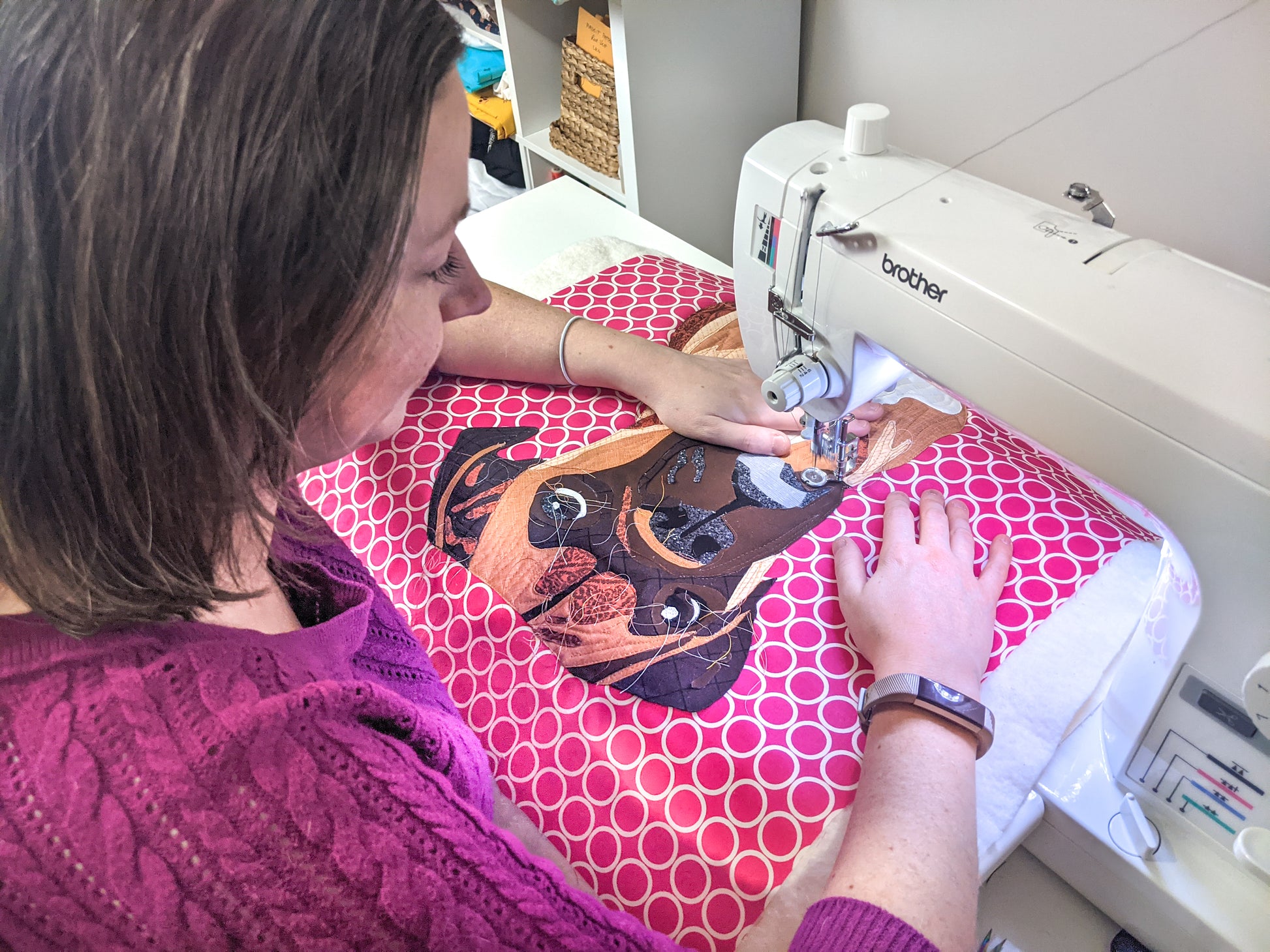 A woman uses Paint By Number Quilts: Dog Portraits from Creative Spark to appliqué a colorful dog face on pink and white fabric, wearing a purple sweater and fitness tracker as she works on her mini quilt.