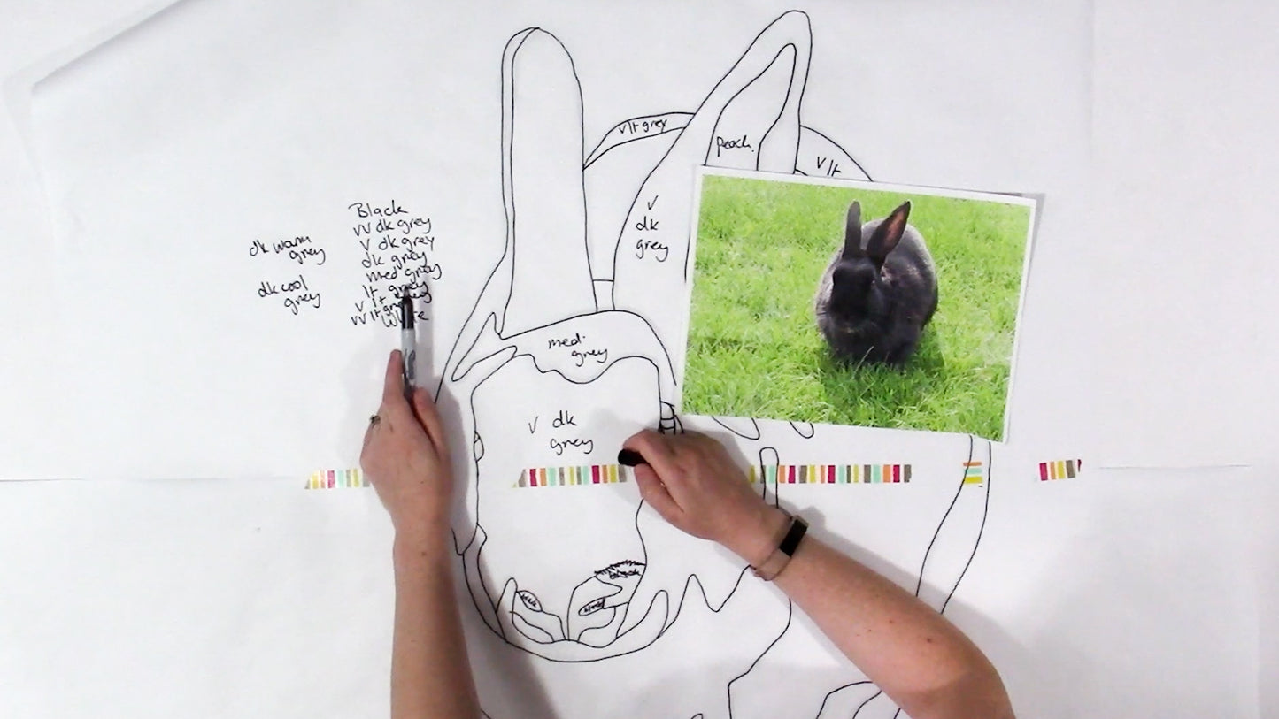 Someone holds a photo of a black rabbit on grass over a large quilt outline, planning their unique wall hanging with notes and colored tape, using the Creative Spark "Design Your Own Paint By Numbers Quilt: Pet Portraits" kit.