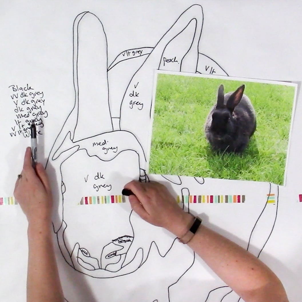 Hands label a line drawing of a rabbit’s head for the “Design Your Own Paint By Numbers Quilt: Pet Portraits” by Creative Spark. A photo of a dark grey rabbit sits atop, with colored stripes outlining the future quilted wall-hanging.