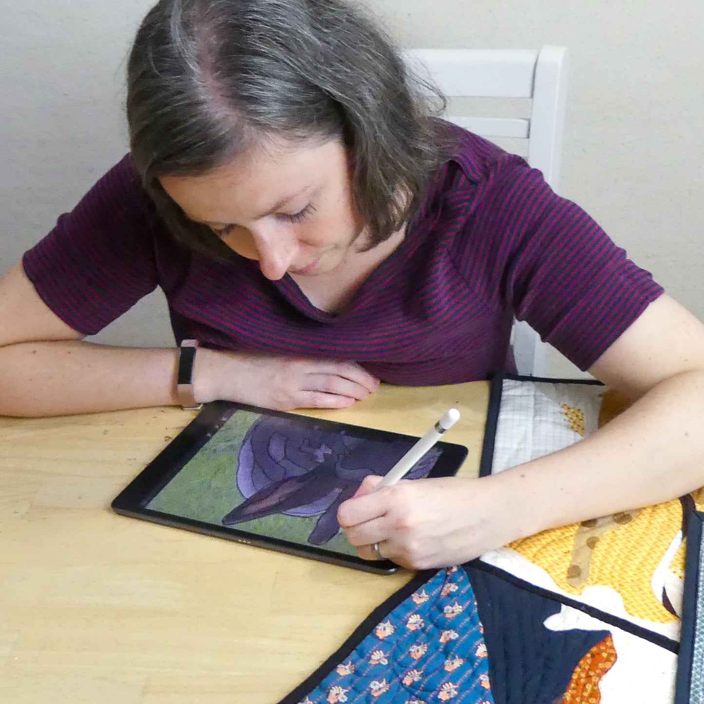 A woman with shoulder-length hair uses a tablet and stylus at a table, next to the "Design Your Own Paint By Numbers Quilt: Pet Portraits" by Creative Spark, focusing intently on her digital artwork.