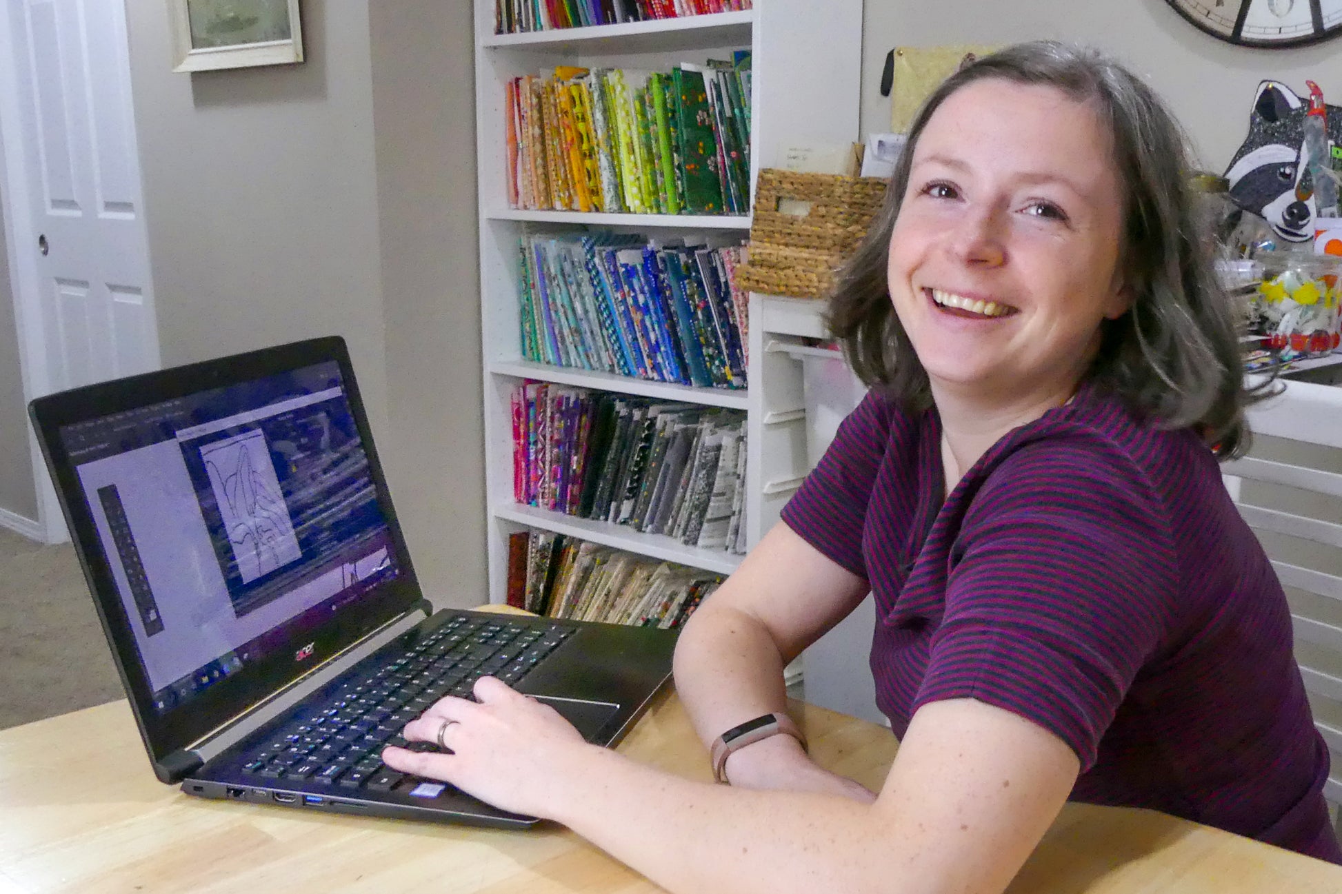 A woman with short brown hair and a striped purple shirt smiles at the camera while using her laptop, with colorful fabric and Creative Spark's "Design Your Own Paint By Numbers Quilt: Pet Portraits" displayed on shelves behind her.
