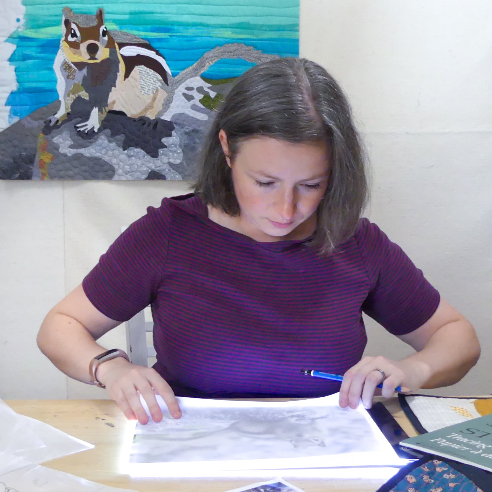 A woman with short brown hair in a purple striped shirt uses a light box at her desk. Behind her, the Creative Spark "Design Your Own Paint By Numbers Quilt: Pet Portraits" adds charm to the space.
