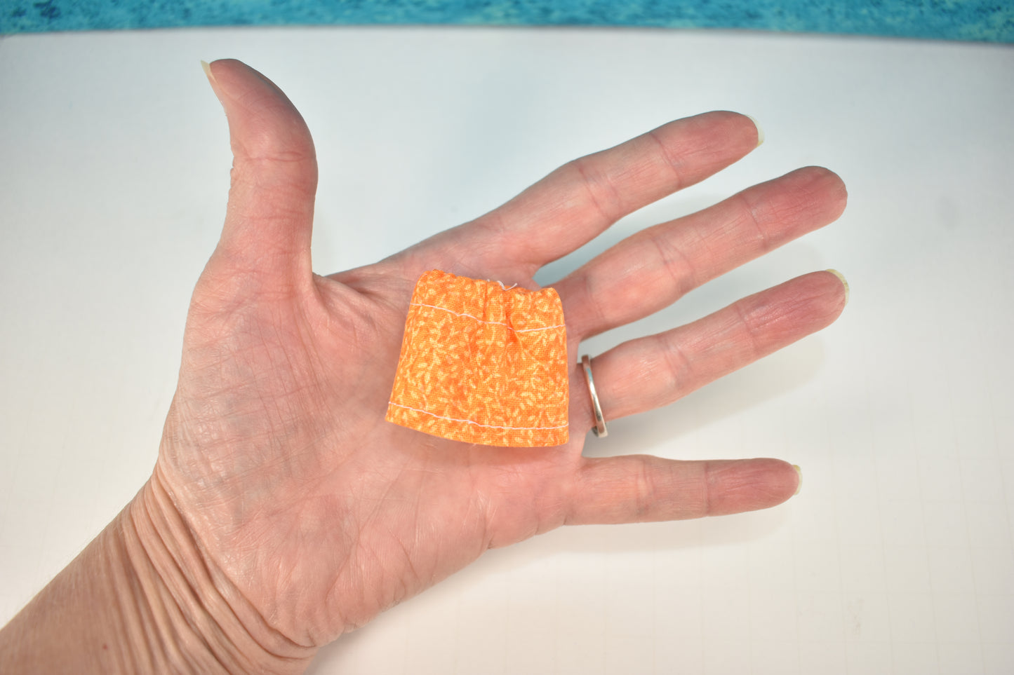 A left hand wearing a silver ring holds a tiny orange patterned doll skirt, illustrating sewing tips from Creative Spark's "How to Alter Doll Clothes Patterns," set against a white background with a blue edge.