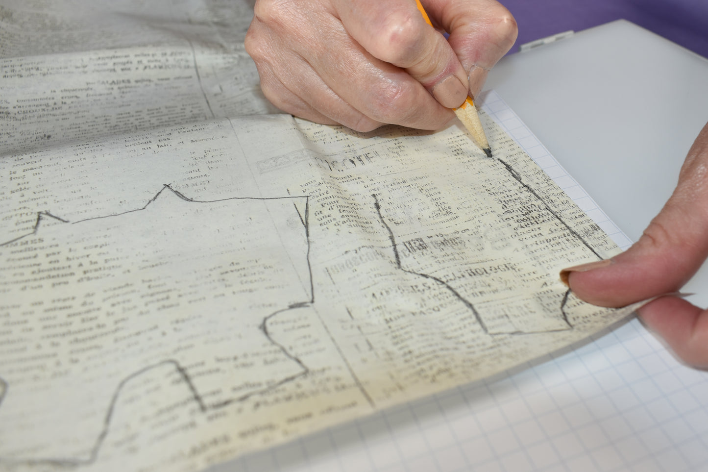 Using Creative Spark's "How to Alter Doll Clothes Patterns," someone traces a graph outline onto old newspaper with a pencil, holding it steady as the paper rests on graph paper—perfect for crafting custom doll clothes.