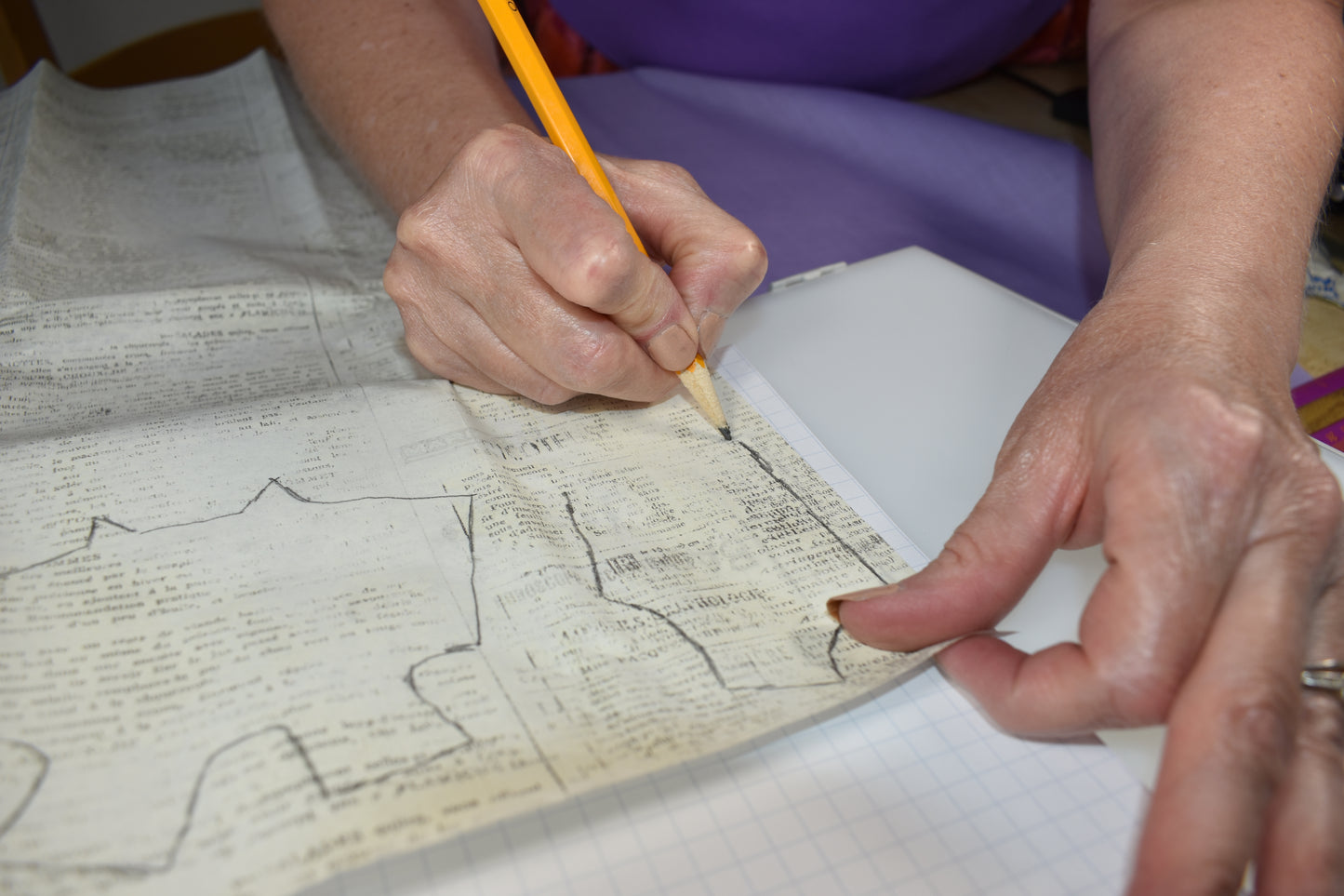A person uses the "How to Alter Doll Clothes Patterns" guide by Creative Spark, tracing printed outlines onto fabric with a pencil on a grid-lined surface, adjusting sewing patterns for custom doll clothes by hand.