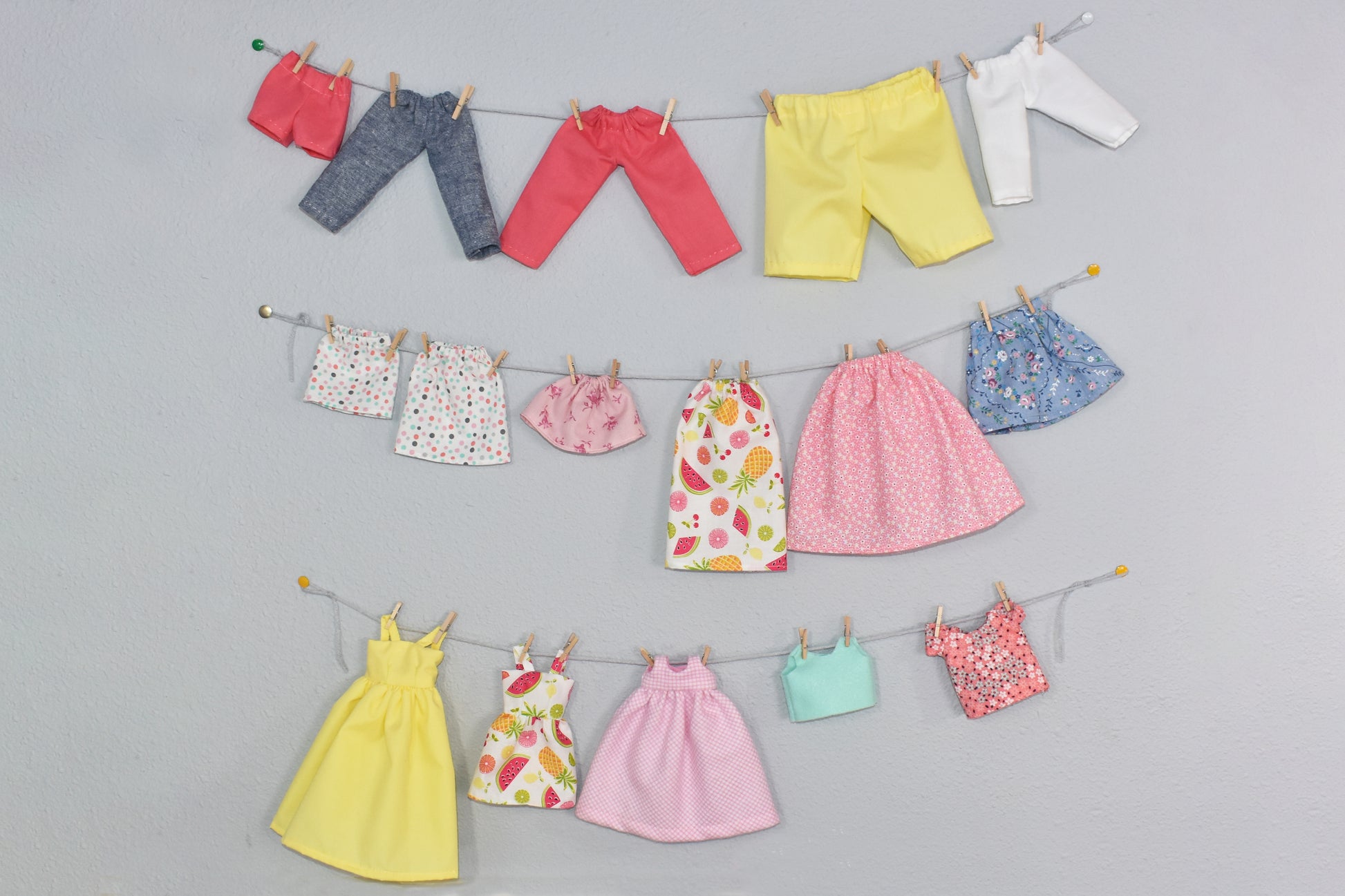 Colorful doll clothes made with Creative Spark’s How to Alter Doll Clothes Patterns hang on three strings with mini clothespins against a light gray wall—showcasing pants, skirts, tops, and dresses in vibrant colors and patterns.