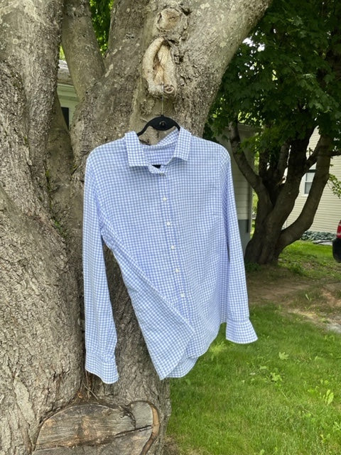 The Creative Spark Stress-Free Shirt-Making features a blue and white checkered long-sleeve shirt, expertly sewn, displayed on a black hanger from a tree branch with green grass and trees in the background.
