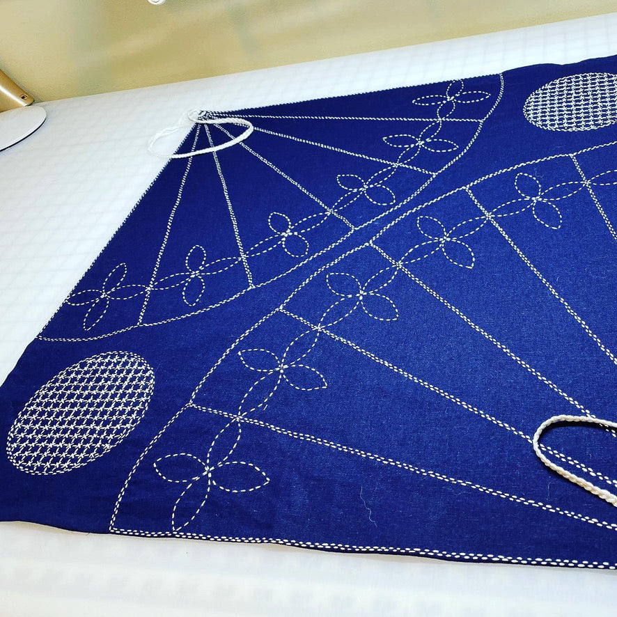 The Creative Spark Make Your Own Furoshiki: Sashiko Embroidered Wrapping Cloth is a blue triangle with white sashiko floral and geometric stitches, corner rope loops, and lays flat on a white checkered surface—an eco-friendly wrap option.