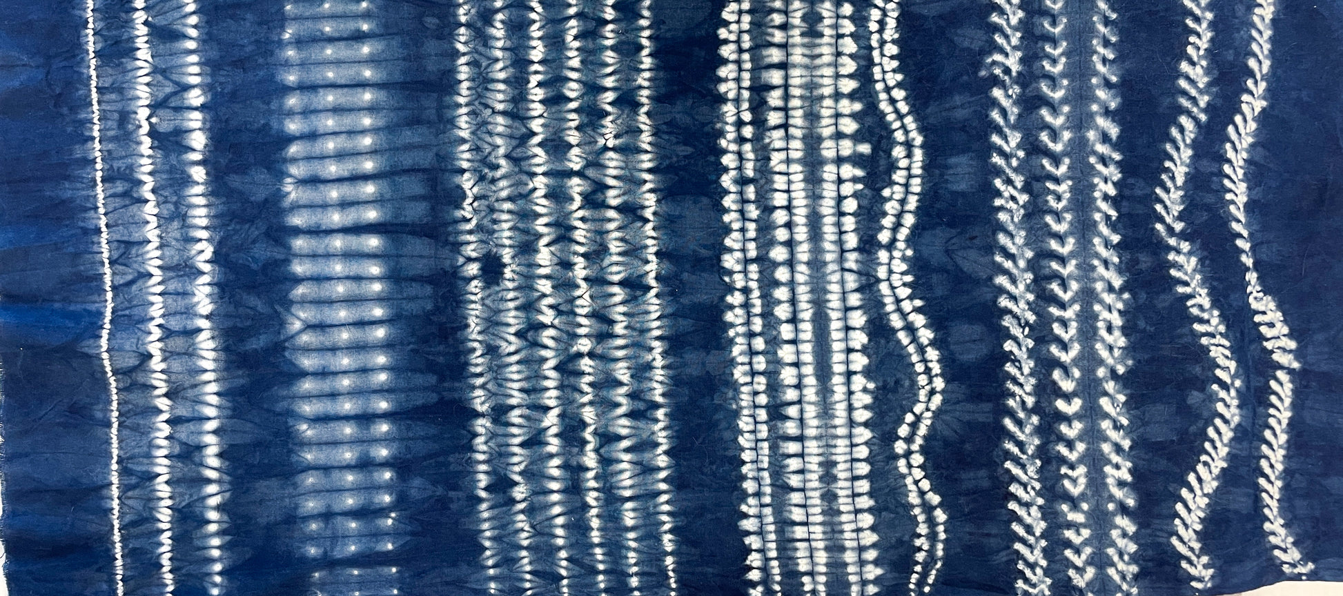 Stitch Resist for Shibori by Creative Spark features blue fabric with vertical white tie-dye patterns—irregular, wavy lines on an indigo background. White lines range from fine dots to broad bands, created using traditional shibori stitch resist techniques.