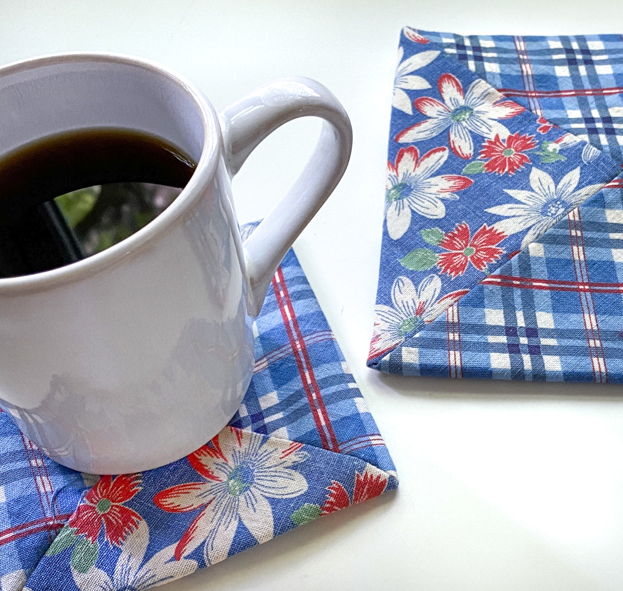 A white coffee mug with black coffee sits on a Creative Spark Feedsack Primer blue plaid coaster with red and white flowers; a matching coaster from the same set lies nearby on the white surface.