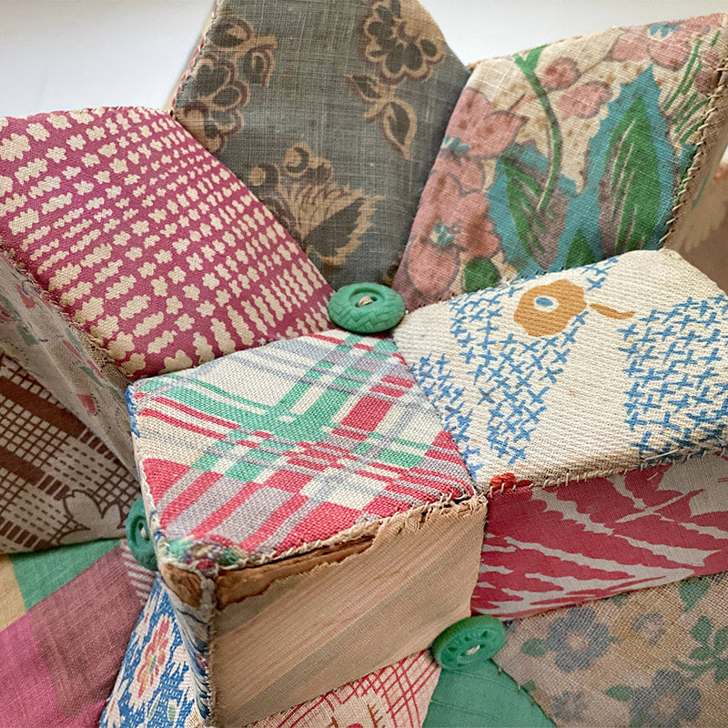 A close-up of Creative Spark's Feedsack Primer shows cubes crafted from vintage-style patterned fabric with green buttons at intersections; visible wear and frayed edges highlight its rich, practical history.