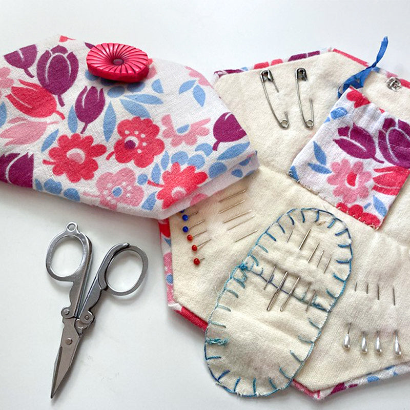 The Creative Spark Feedsack Primer kit is open, showing neatly arranged needles, pins, and safety pins. Scissors and a matching closed pouch with a red button sit nearby on a white surface—ideal for any sewing project.