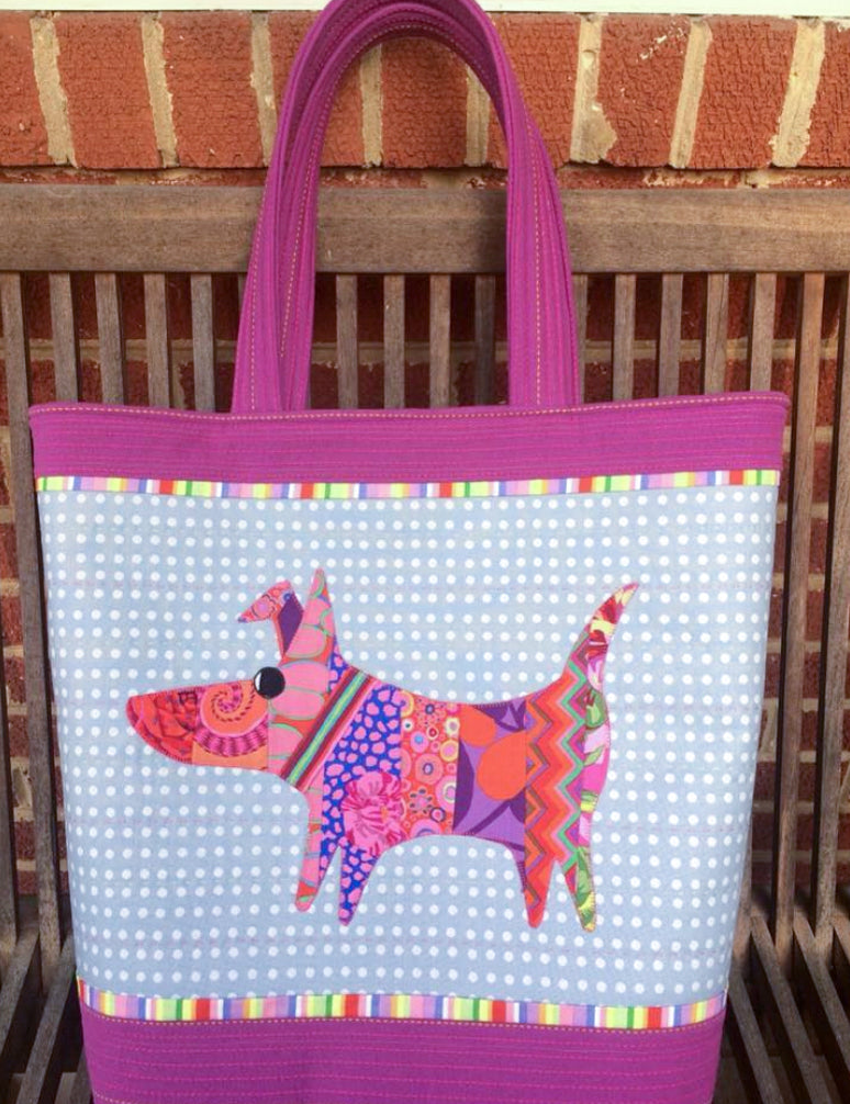 The Creative Spark "Wonky Piecing & Easy Appliqué Mini Mod Dog" tote bag features a patchwork dog on a blue polka dot background with rainbow borders and purple handles, displayed on a wooden bench.
