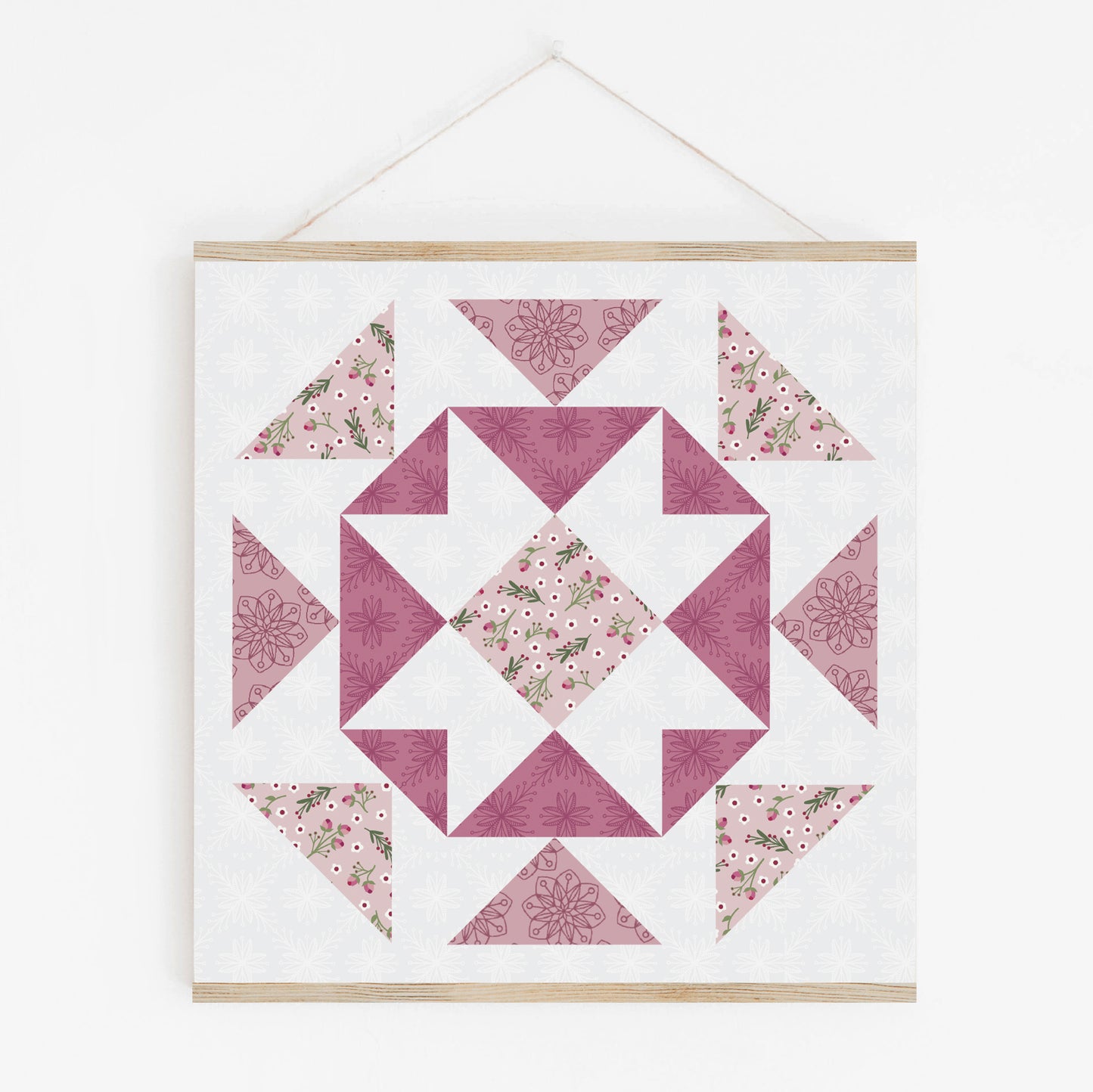 The Yuletide Sampler Block-of-the-Month Quilt by Creative Spark showcases geometric pink, purple, and white floral and star patterns—perfect for your next quilt along—framed in a light wooden holder on a white wall.