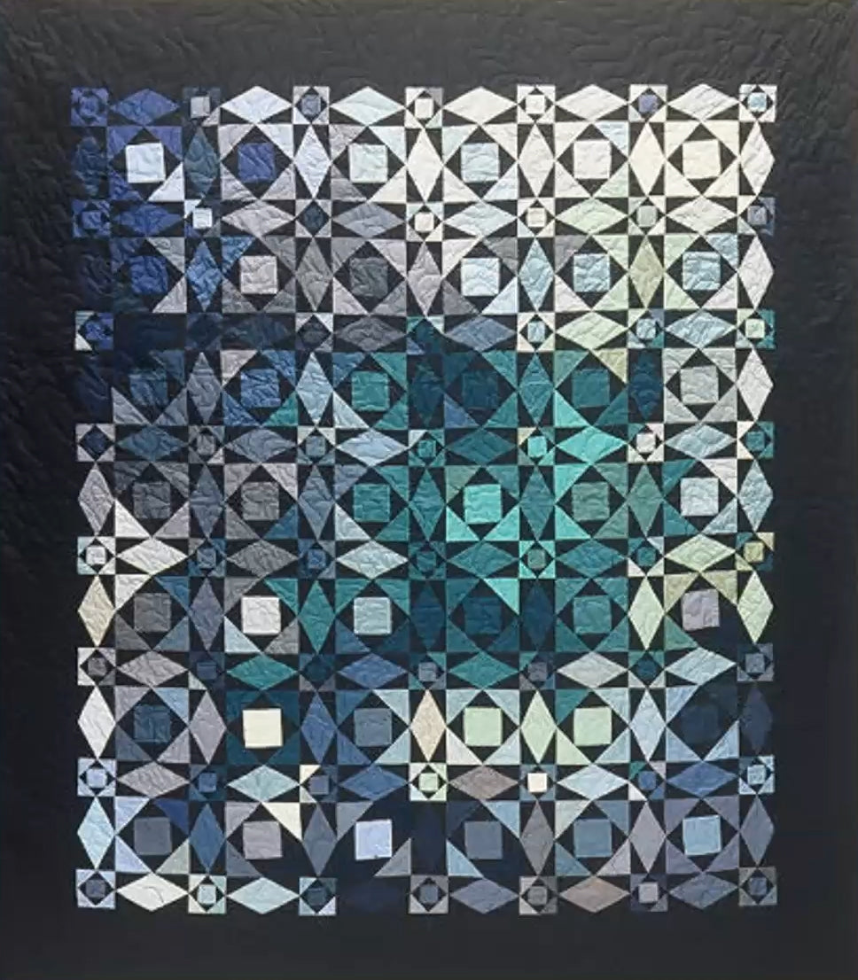 The Creative Spark "What’s Up With Color?" quilt features intricate geometric patterns in blue, teal, gray, and white, arranged in a grid with colors transitioning from light at the top left to dark at the bottom right.