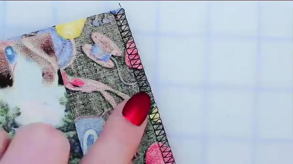 A hand with red nail polish points to a colorful, overlocked fabric edge, highlighting expert seam skills taught in "12 Essential Seams for Great Sewing" from Creative Spark, on a white grid-patterned background.
