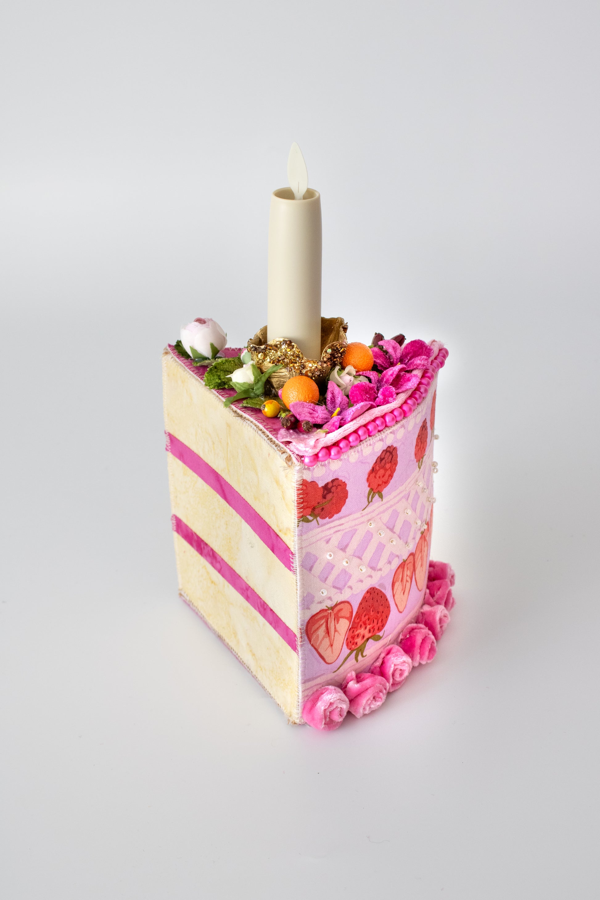 The Piece of Cake Candleholder: A DIY Tabletopper by Creative Spark is a whimsical cake-shaped holder with pink icing, strawberries, tiny fruits, and flowers, topped with a cream-colored candle against a plain white background.